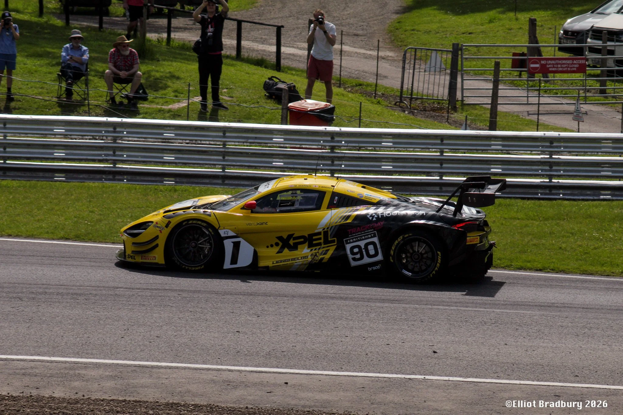 A yellow race car driving on a racetrack with spectators and photographers watching from the grass beyond a barrier fence.
