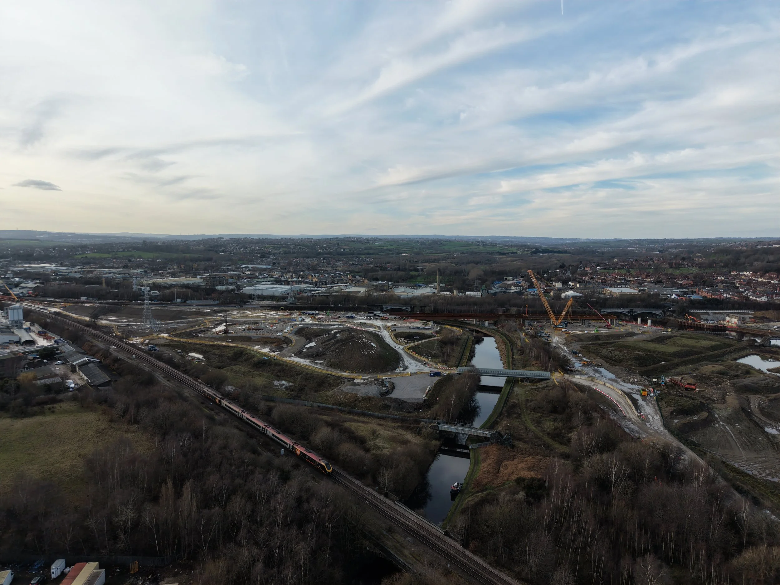 A vast landscape view showing a city or town with buildings, roads, and construction cranes. In the foreground, a railway track with a passing train is visible. Several bodies of water are interspersed throughout the area. The sky is partly cloudy wi