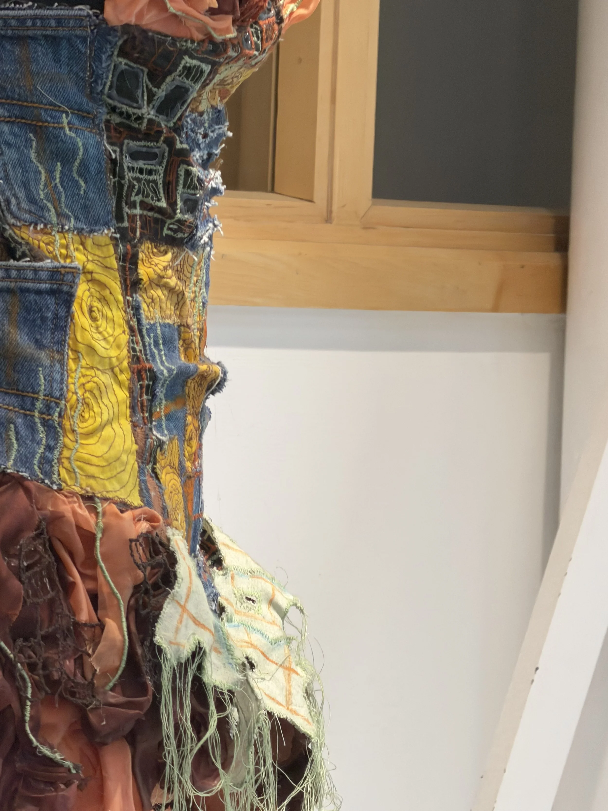 Close-up of a multicolored, heavily distressed and patched fabric with various textures and stitching details, set against a background of a wooden door frame and white wall.