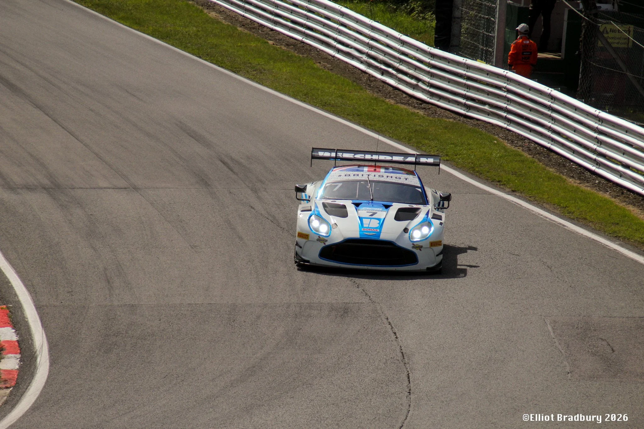 A race car with the number 7 on the front speeds along a racing track, with a safety barrier on the side and a person in an orange jumpsuit and white cap standing near a gate in the background.