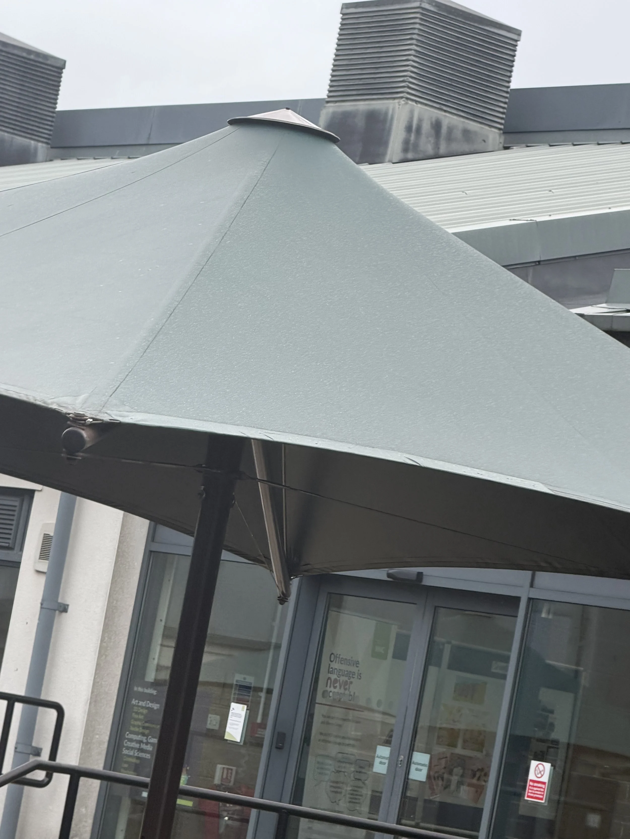 The image shows the entrance of a building with glass doors, with a large grey metal overhang or awning above the doorway, supported by black metal posts. Behind the glass doors, signage for the College of Computing, Computing & Social Sciences is vi