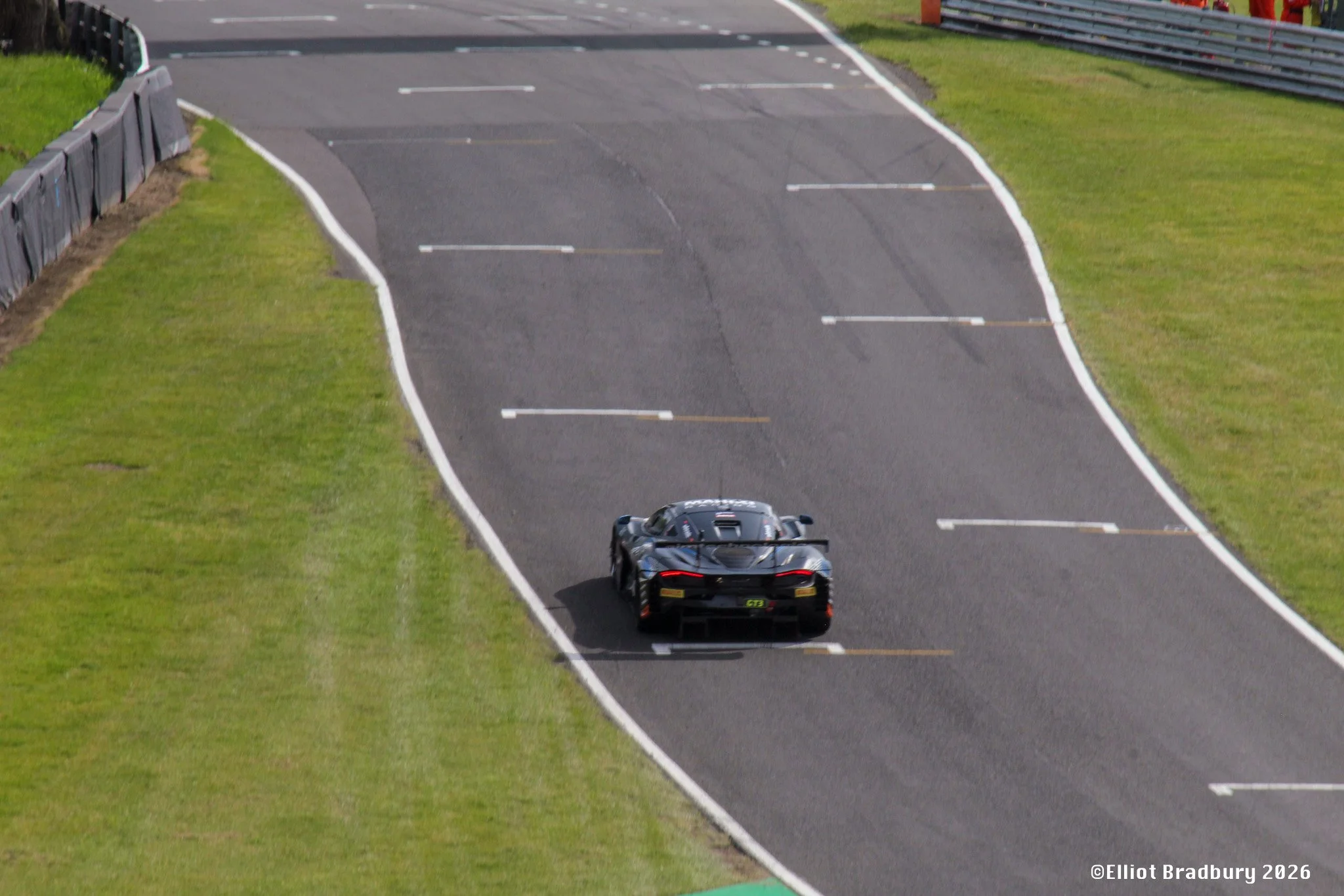A black sports car driving on a race track with a green grass border on both sides and barriers on the left side, approaching a bend in the track.