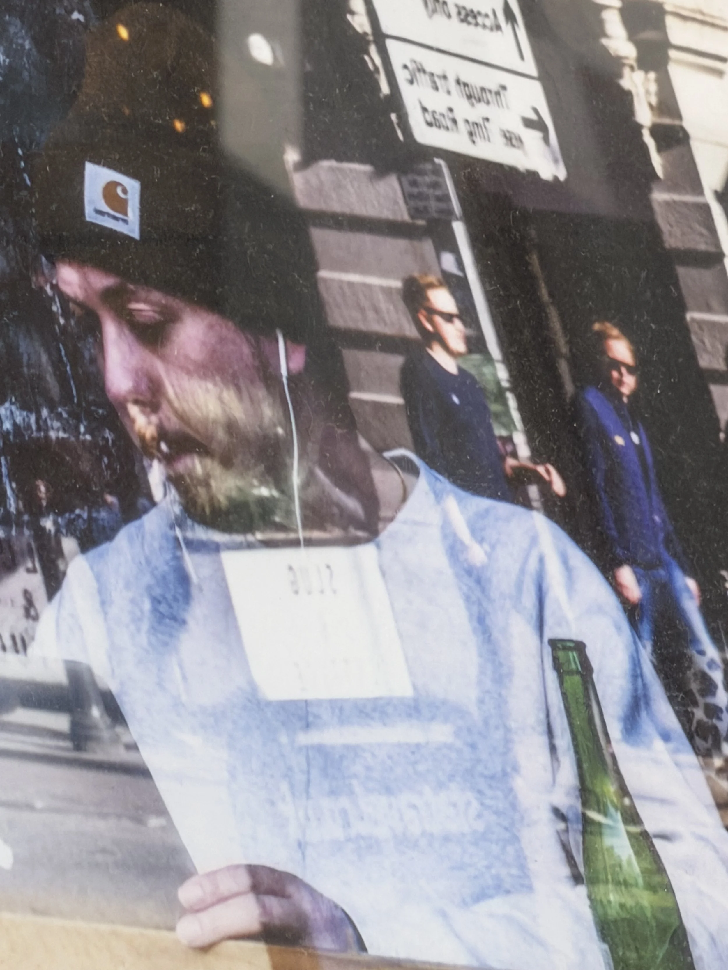 Close-up of a man with glasses and a beard wearing a light gray jacket, looking down at a piece of paper or document. In the background, there are people walking and a street sign in a different language.