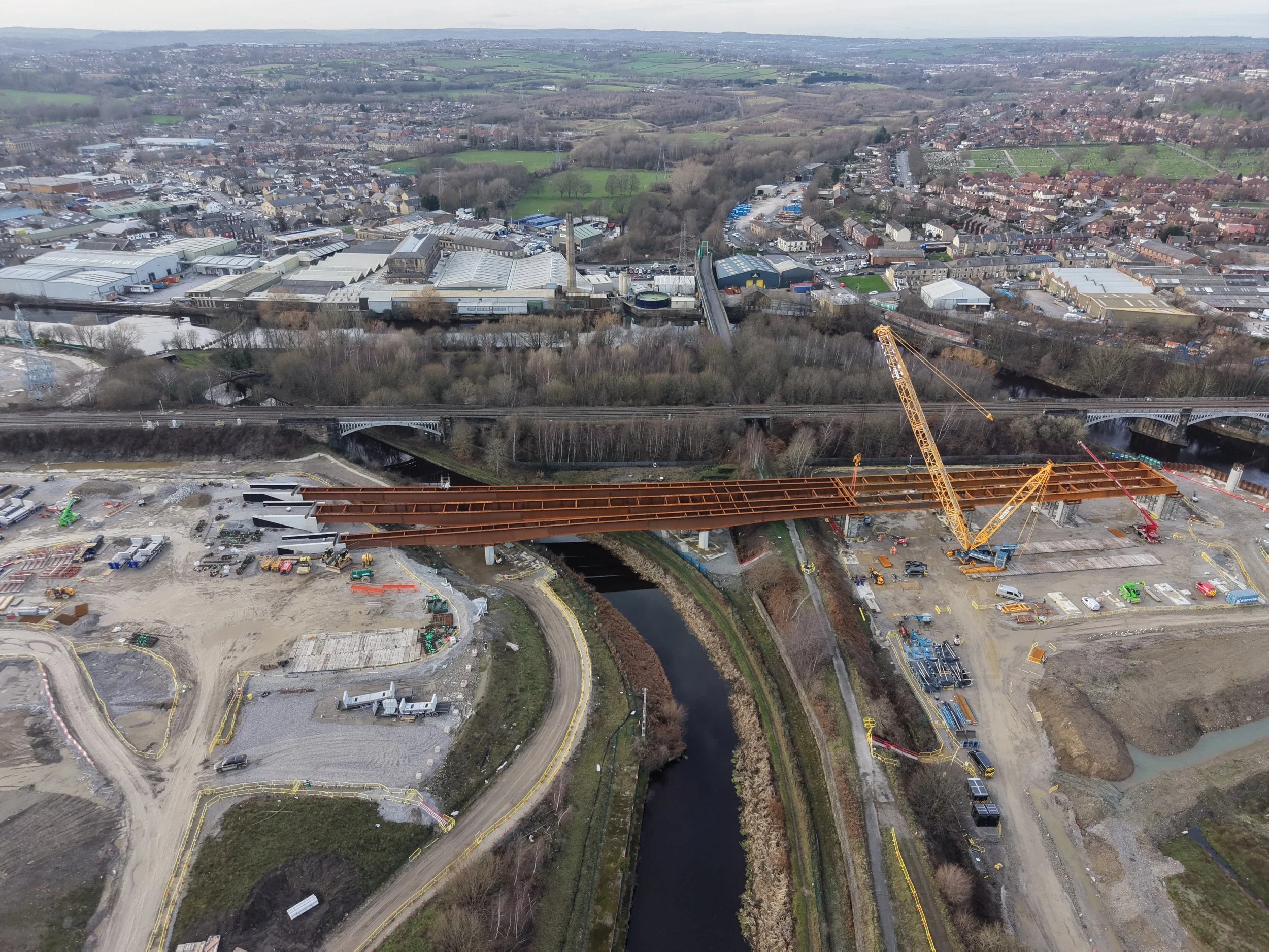 An aerial view of a construction site with a partially built bridge spanning a river, surrounded by residential and industrial buildings, roads, and green fields.