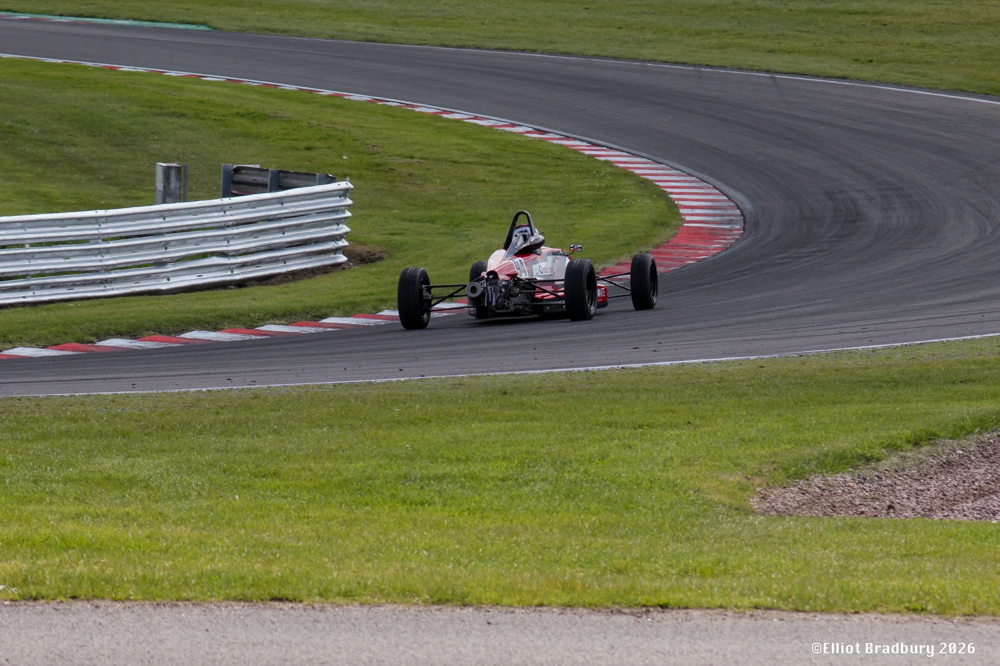 A race car on a curving asphalt track with red and white curbs, a grassy infield, and a metal barrier on the side.