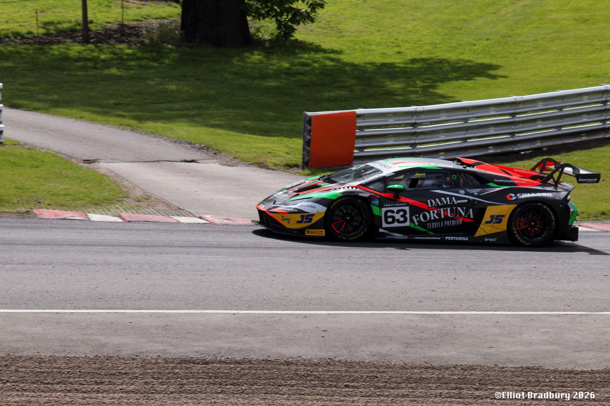 A racing car with the number 63 and sponsorship logos speeding on a race track, with grass and safety barriers in the background.