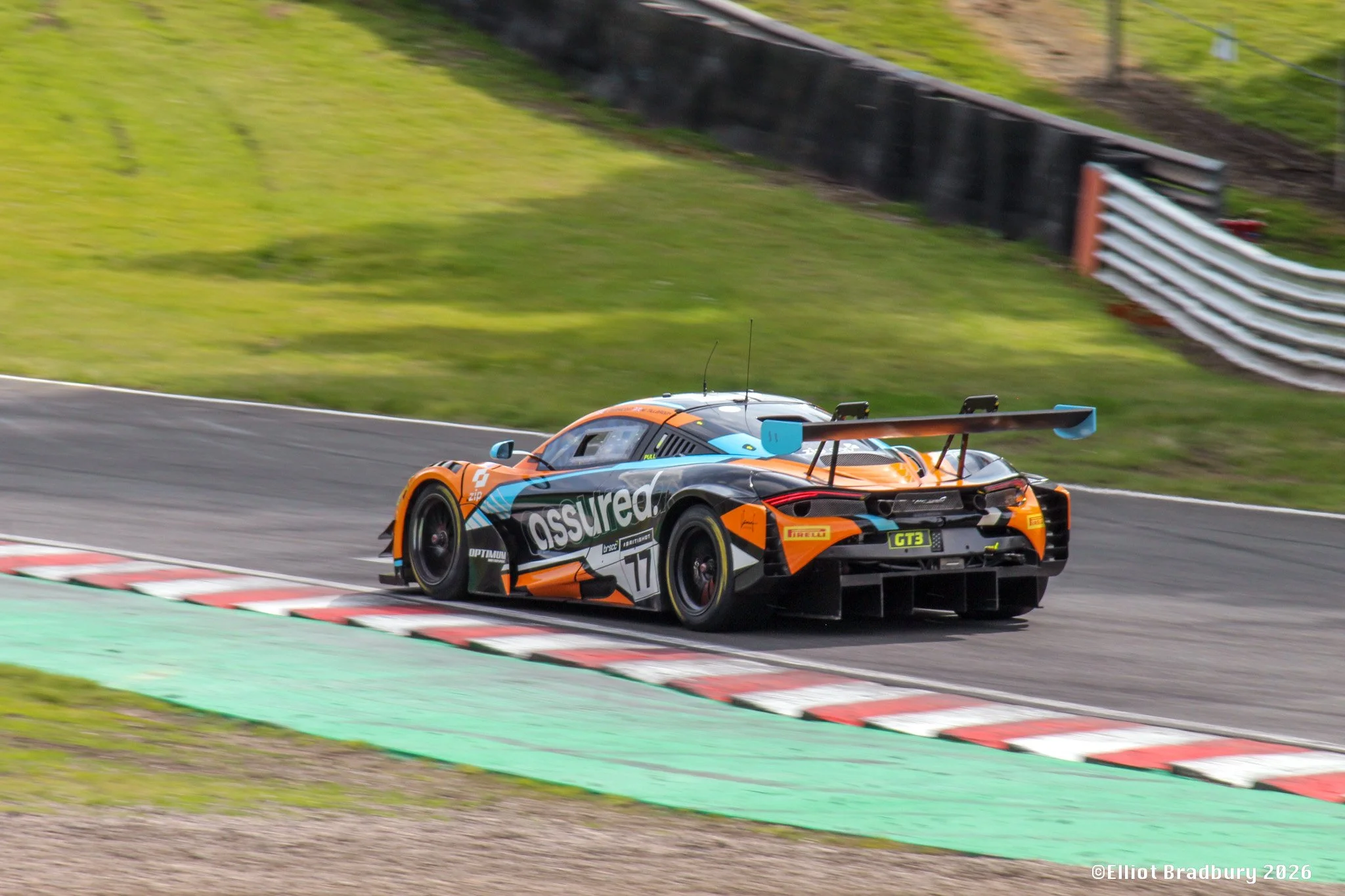 A race car on a racing track, leaning into a turn with the curb and grass visible on the side. The race car is orange and black with various sponsor logos.