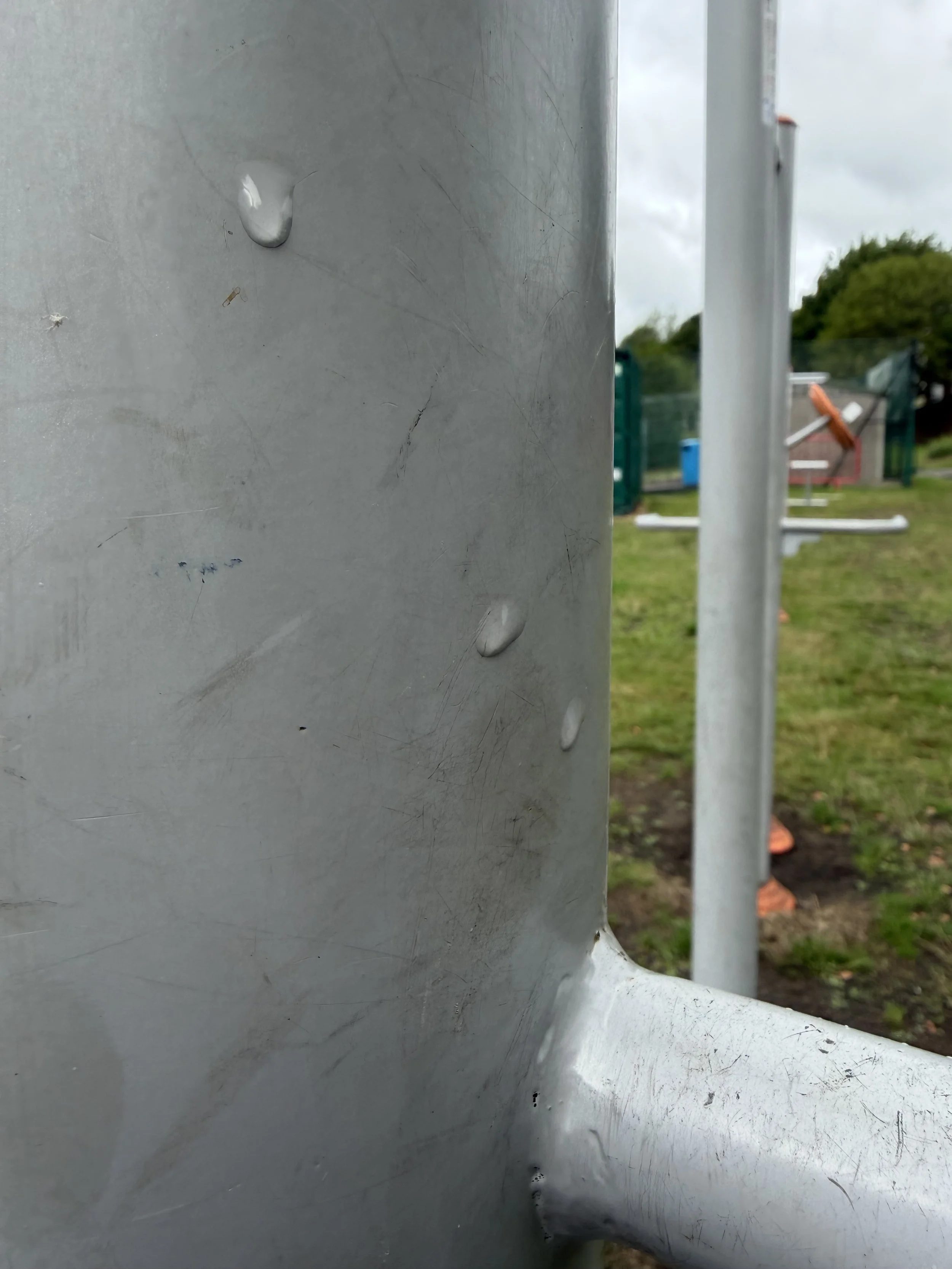 Close-up photograph of a metal structure with water droplets on its surface, outdoors with a background of grassy area, trees, and playground equipment.