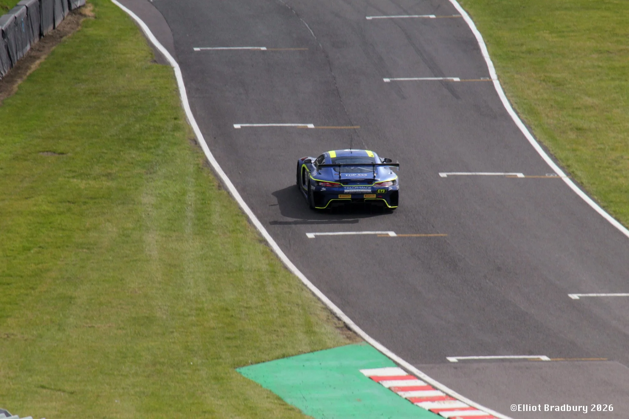 A racing car on a paved race track, with grassy areas on either side, approaching a corner with curved white boundary lines and a green and red striped curb.