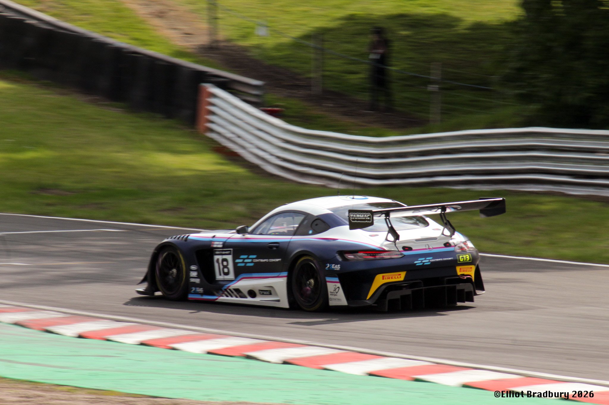 A race car with the number 18 on the side, painted in black, blue, and white, driving on a race track next to green grass and tire barriers with a blurred background.