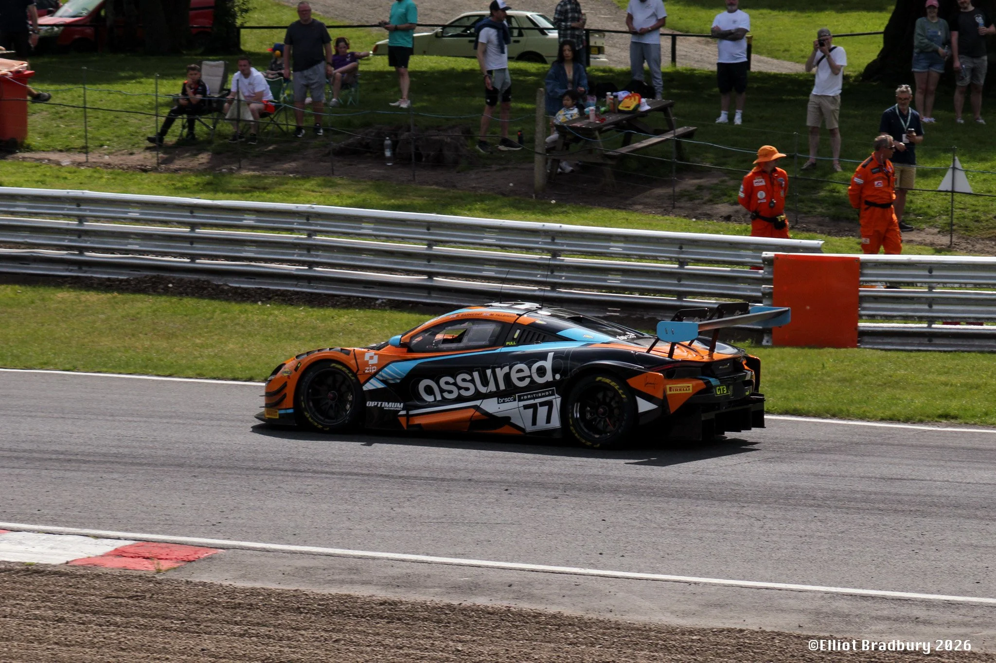 A race car with the word 'assured' on its side speeds along a race track. Spectators and race officials stand behind a safety barrier watching the race, with some taking photos. There are trees, parked cars, and a picnic table in the background.