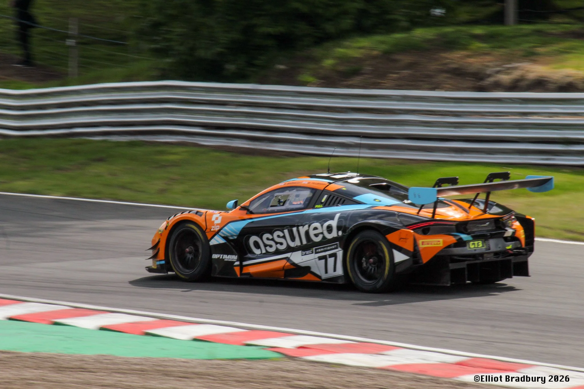 A race car on a track, painted orange and black with sponsor logos, speeding past a metal guardrail and green grass in the background.