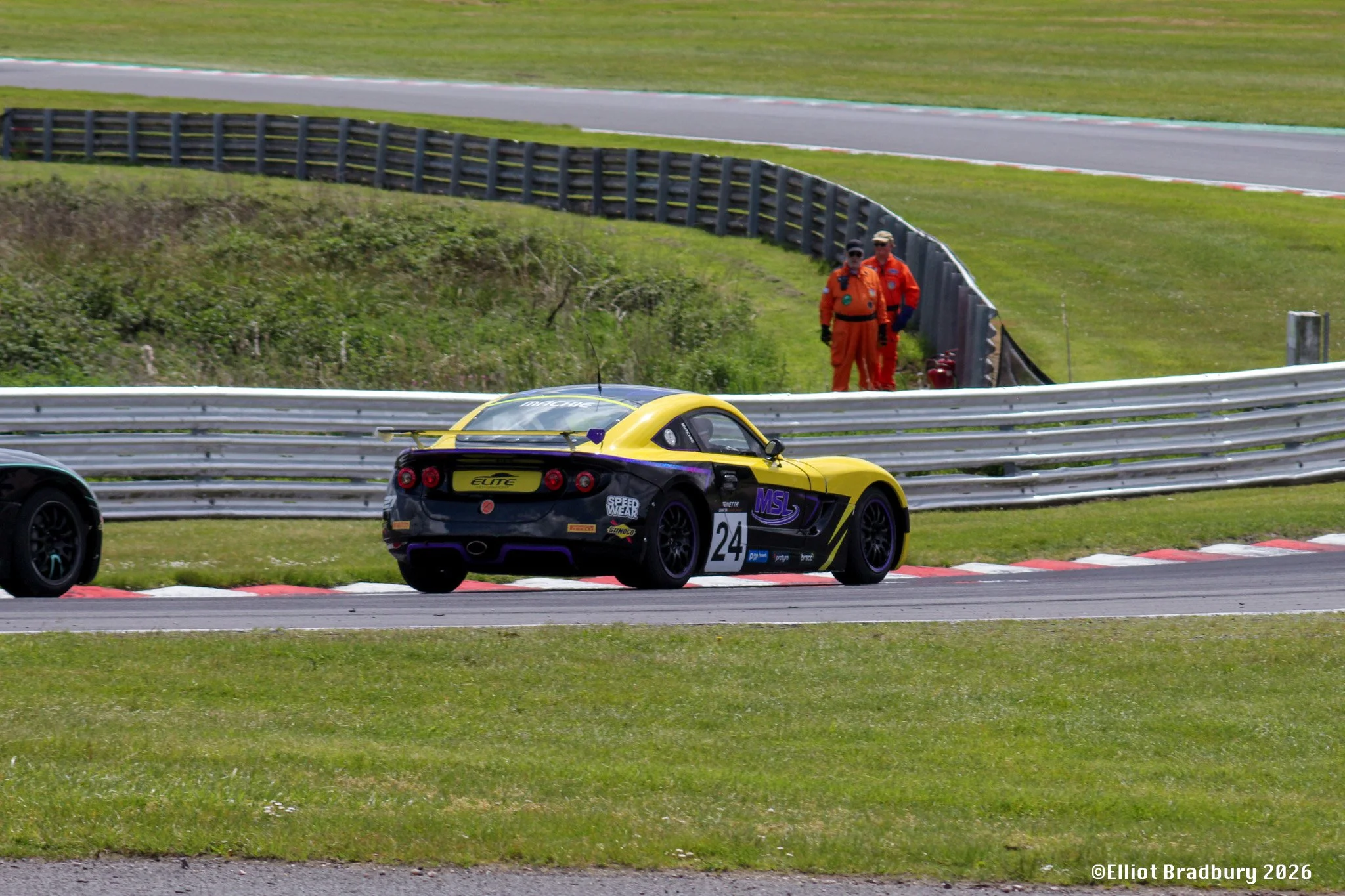 A yellow and black race car on a track, with two race officials in orange uniforms standing nearby on a grassy area, and a safety barrier separating the track from the officials.