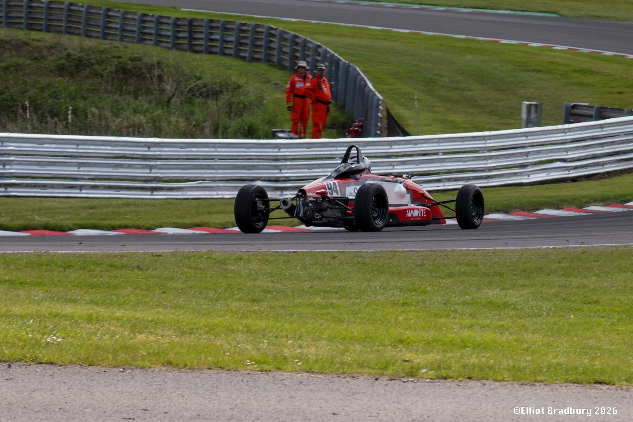 A red and white race car with the number 94 on the side crashing into the grass and barriers on a racetrack, with two rescue workers in orange uniforms standing nearby.
