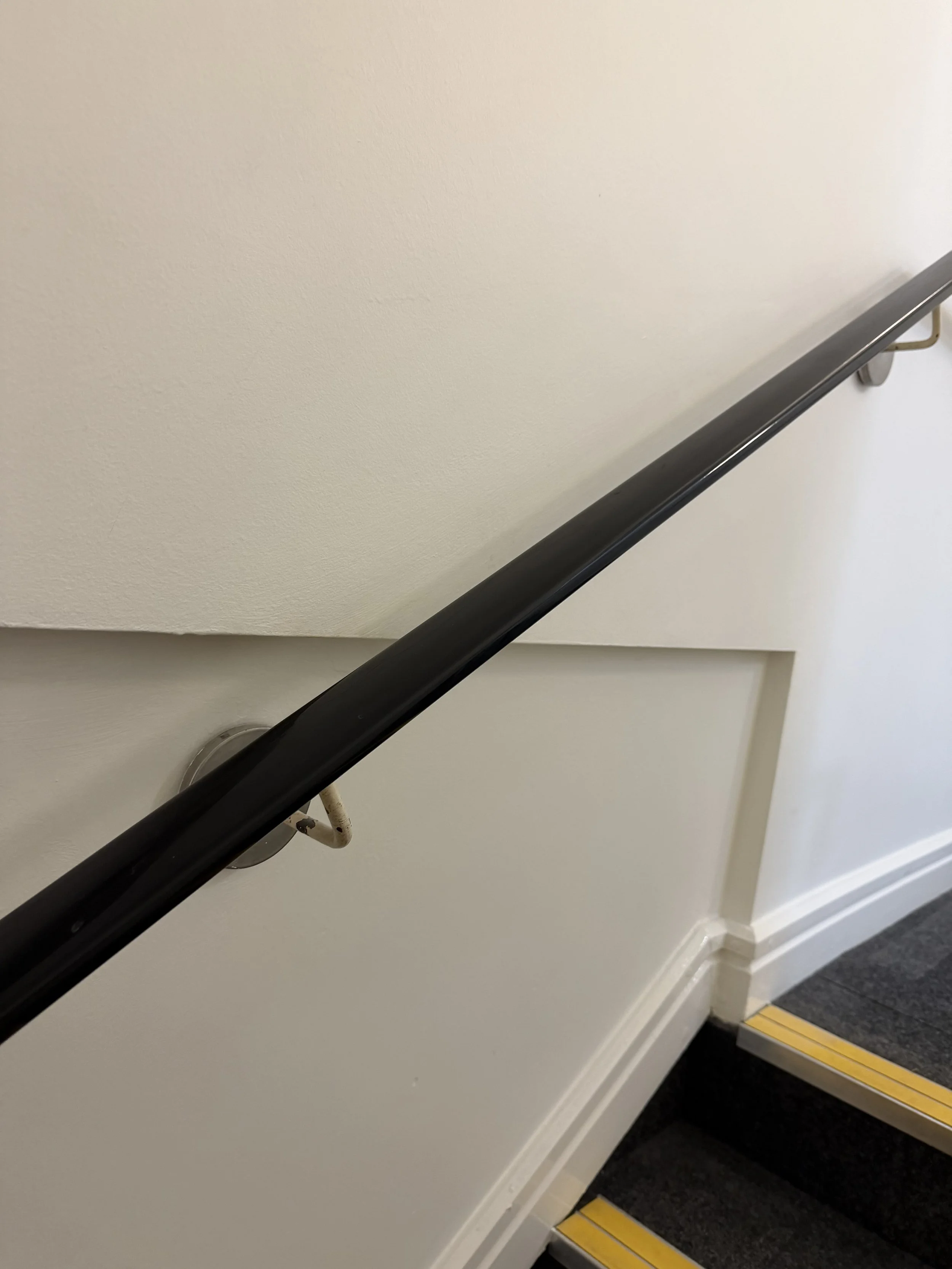 Close-up of a black handrail along a wall, with a yellow stripe on the edge of a step at the bottom right corner.