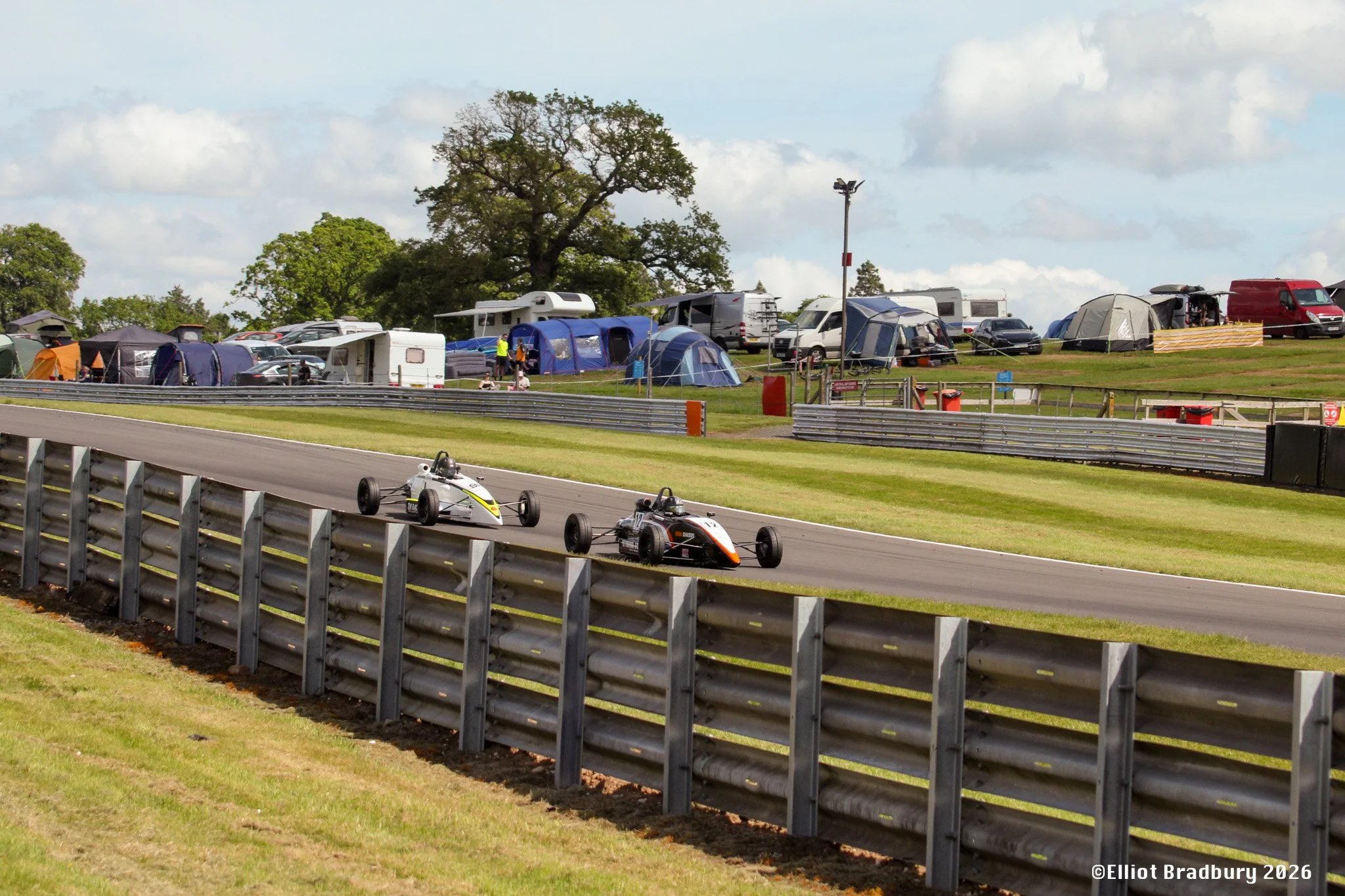 Race cars on a track with tents and cars in the background, spectators and marshals near the tents, and a large tree and cloudy sky overhead.