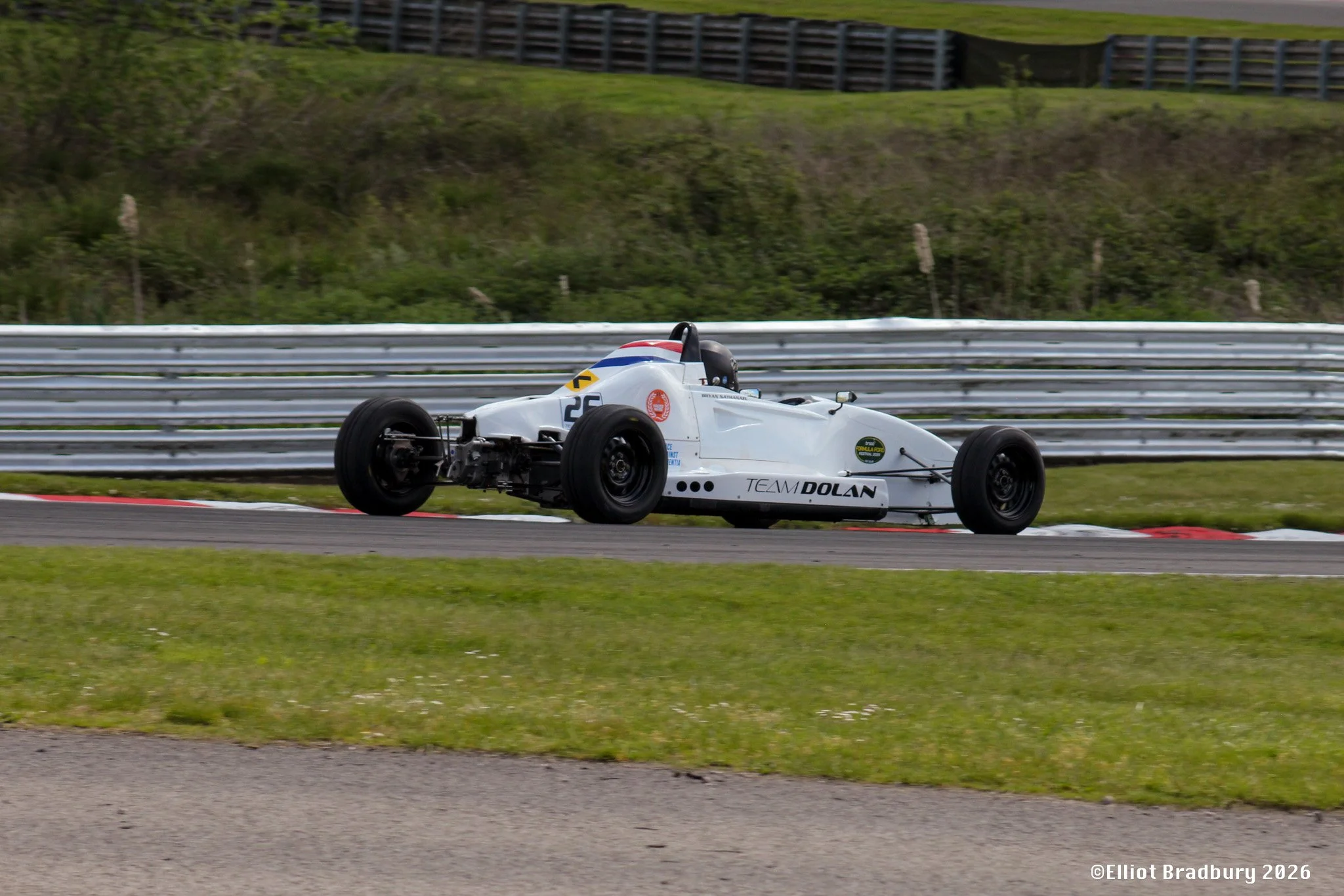 A white Formula race car with black tires and colorful decals on a race track, navigating a curve with green grass and a safety barrier in the background.