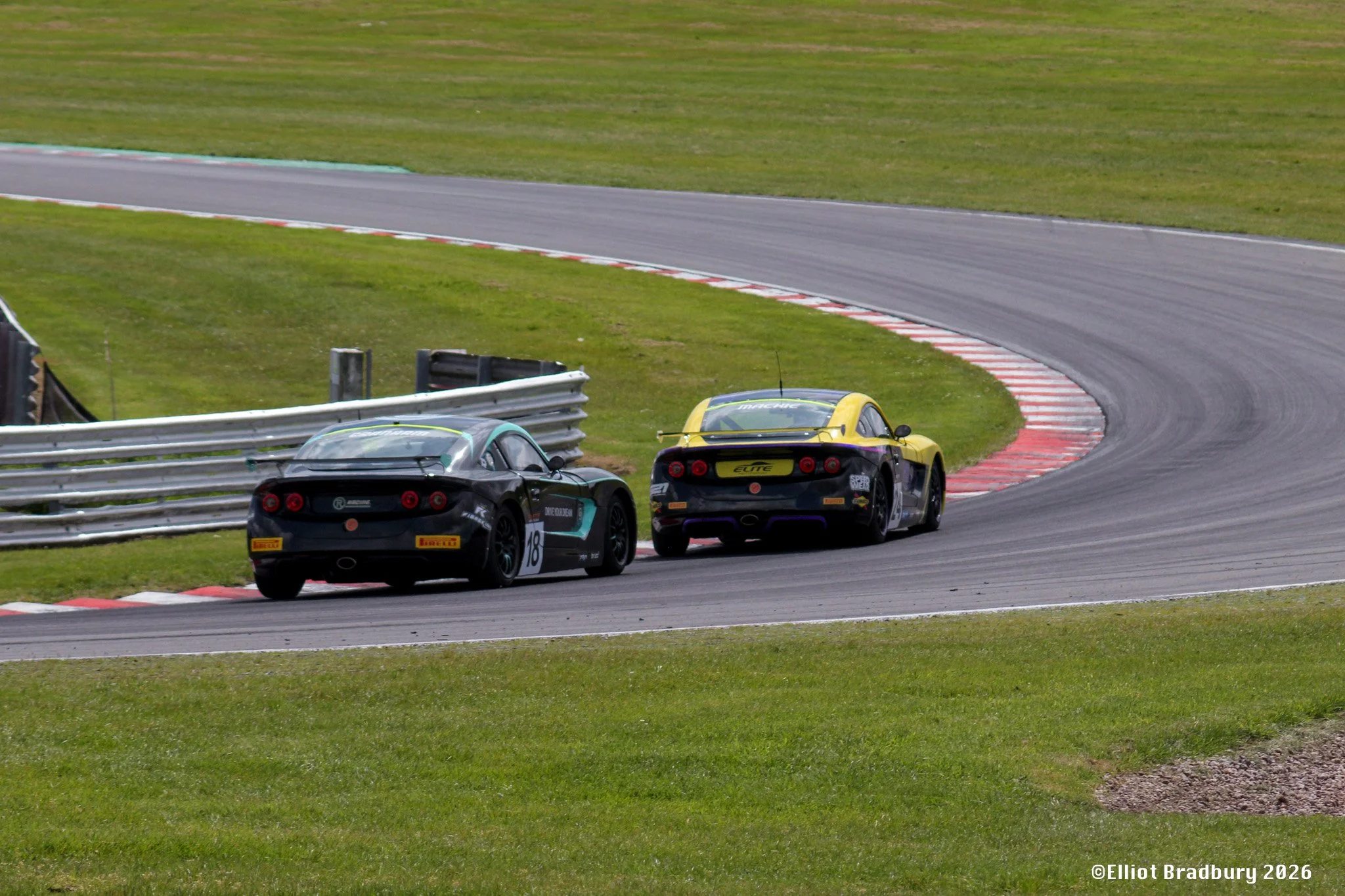 Two race cars on a curved section of a race track, with green grass and a metal guardrail nearby, during daytime.
