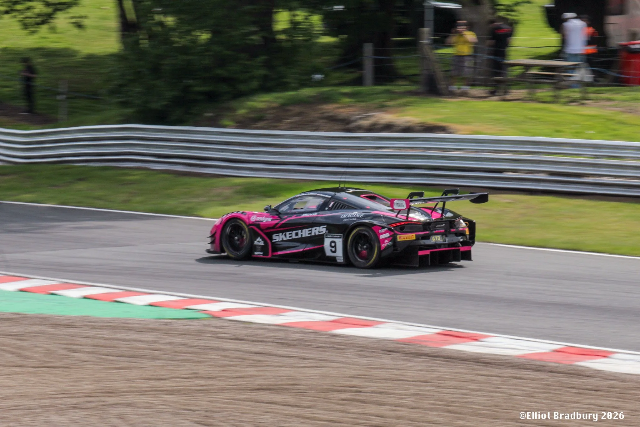 A black and pink race car speeding along a racetrack on a curve, with a safety barrier and trees in the background.