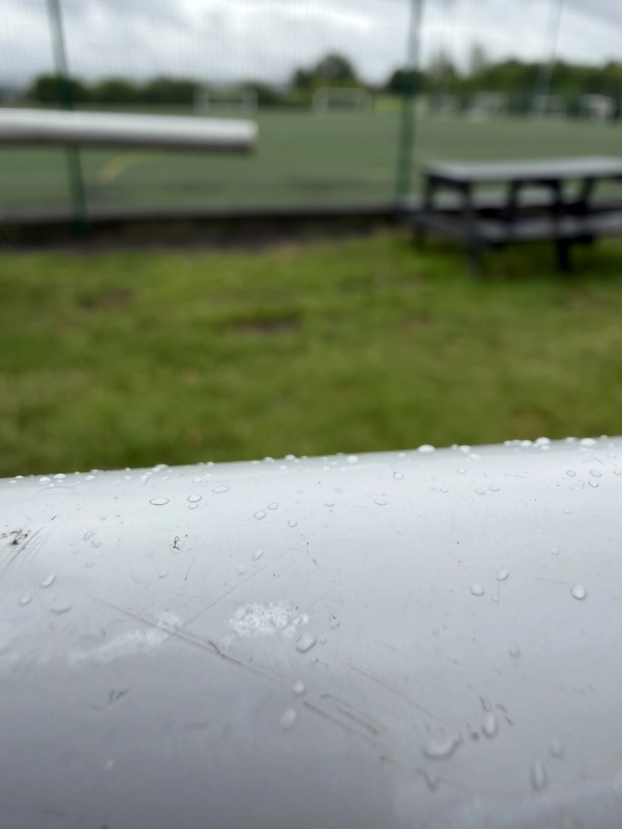 View through a rain-covered window showing blurred outdoor sports field, benches, and trees in the background.