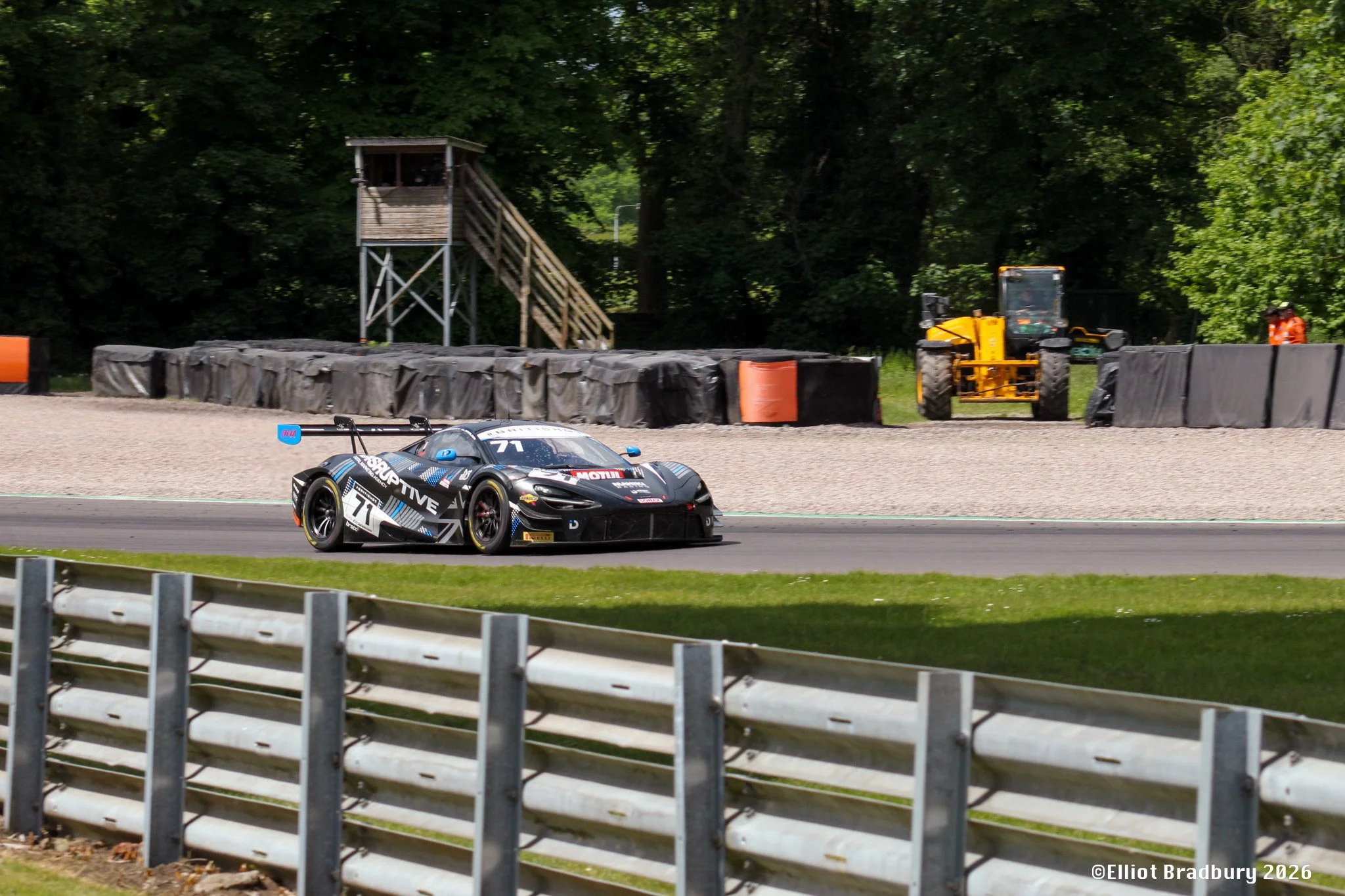 A race car on a track, with trees and a watchtower in the background, along with a construction vehicle and two race officials nearby.