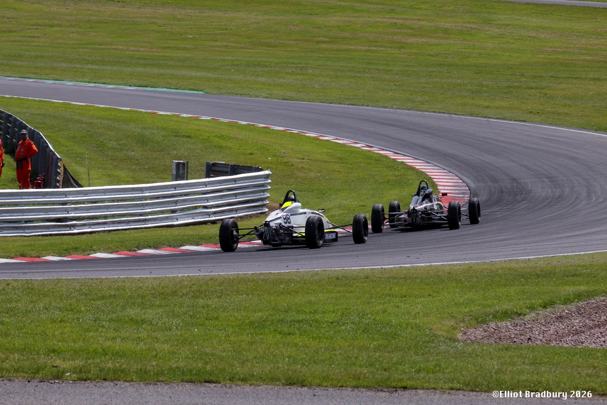 Two formula-style race cars on a racetrack navigating a curve with a grass infield and safety barriers, with a person in red standing near the barriers.