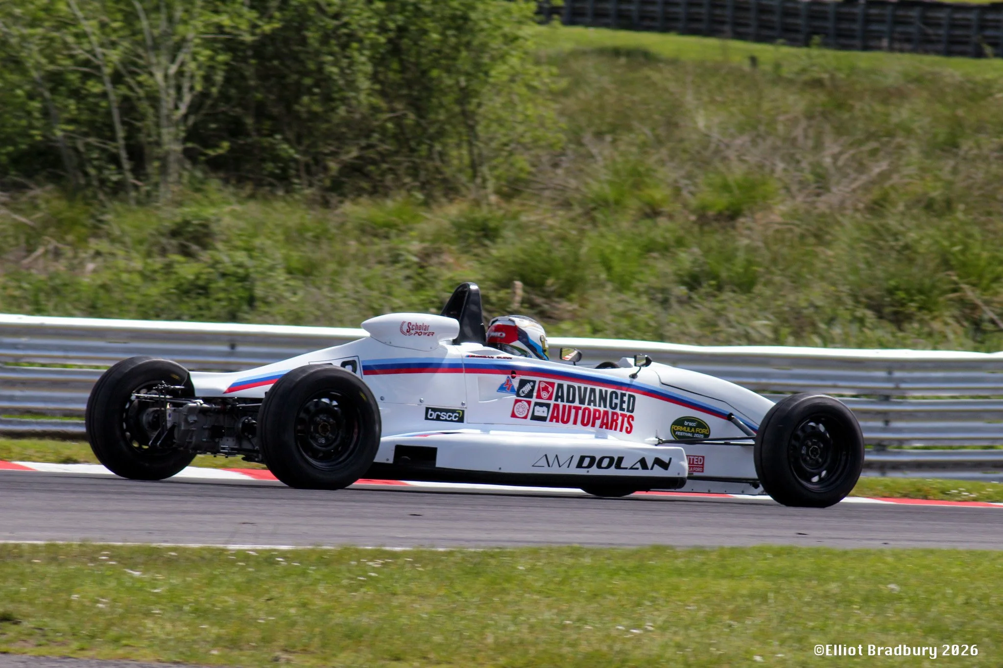 A white race car with various sponsor logos, including 'Advanced Autoparts' and 'Dolan,' is speeding on a racetrack with a grassy area and green trees in the background.