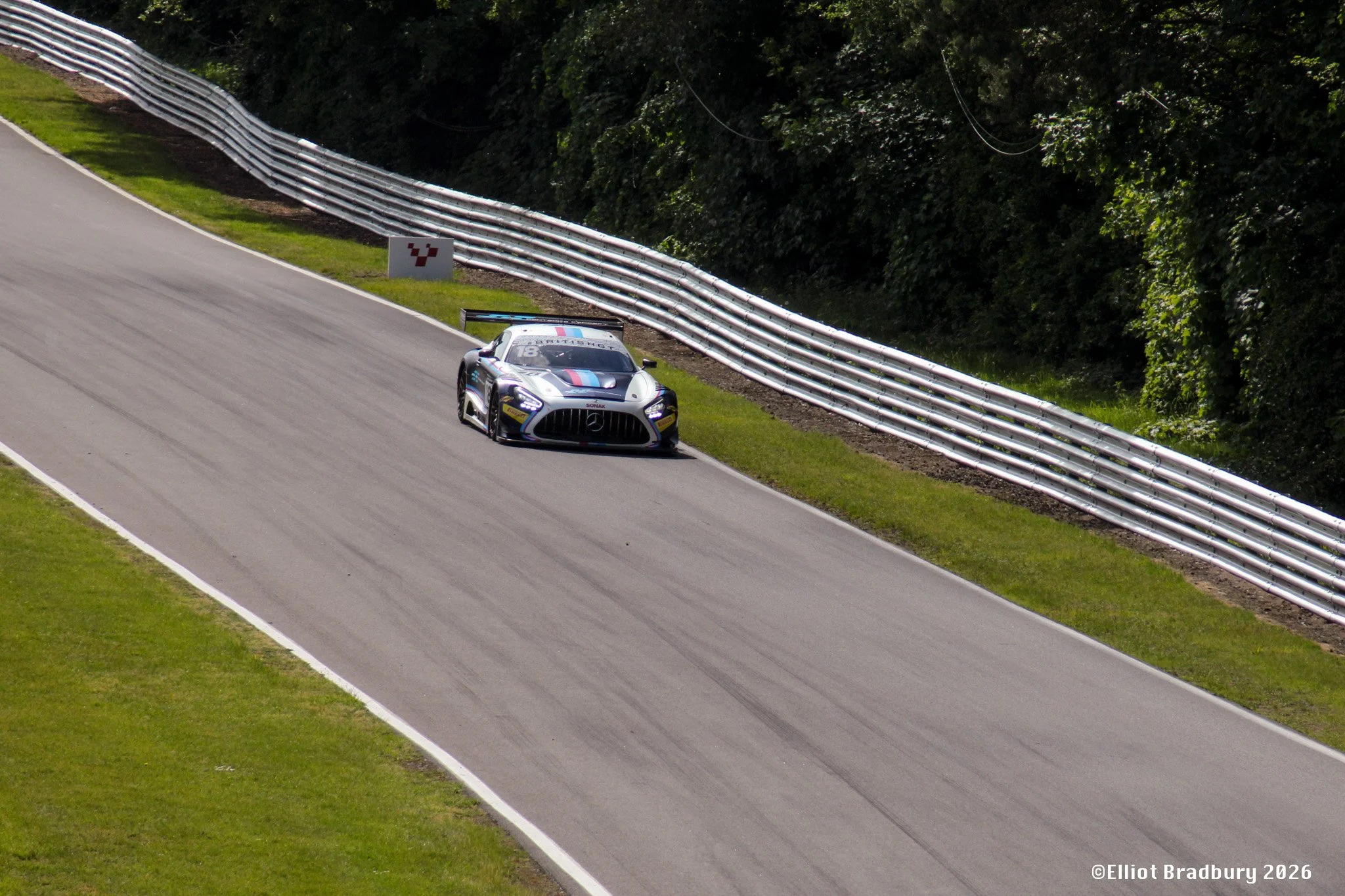 A high-performance race car driving on a curving race track with metal barriers and dense green trees in the background.