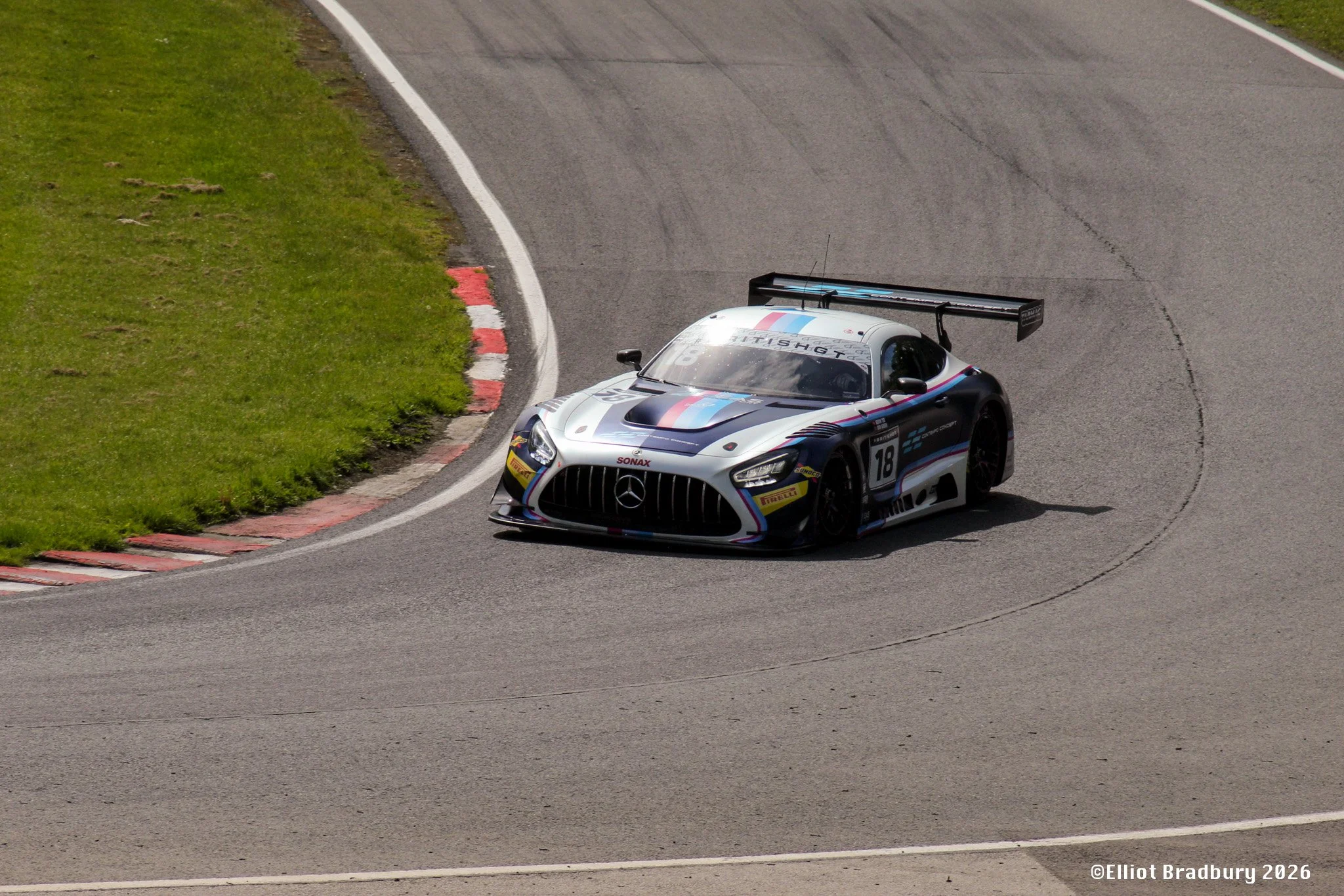 A silver race car with a black, red, and blue stripe design on a curved race track next to green grass. The car has a large rear spoiler and various sponsor decals.