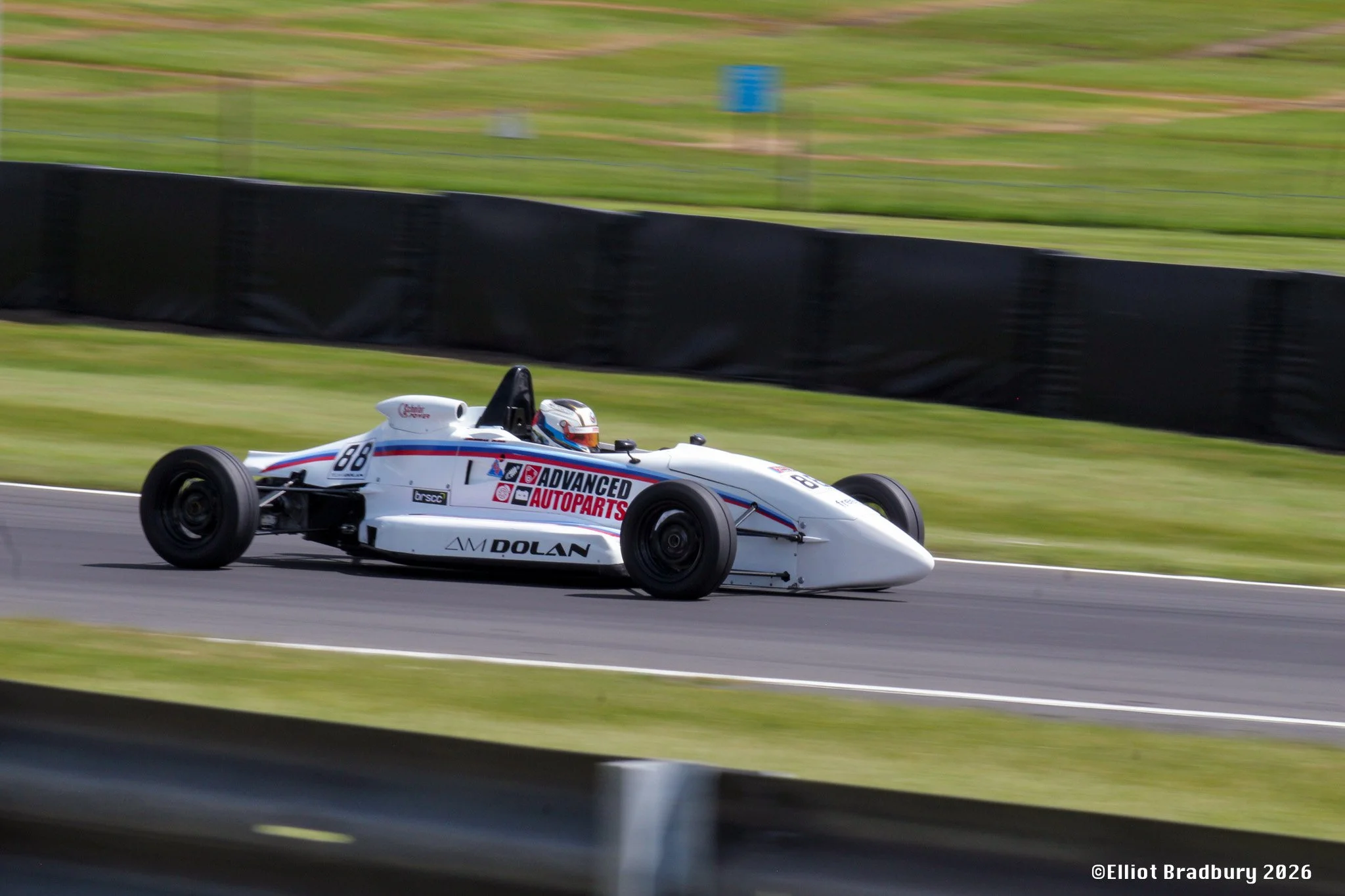 A white race car with sponsorship decals speeding on a racetrack, with motion blur in the background and black barriers along the track.
