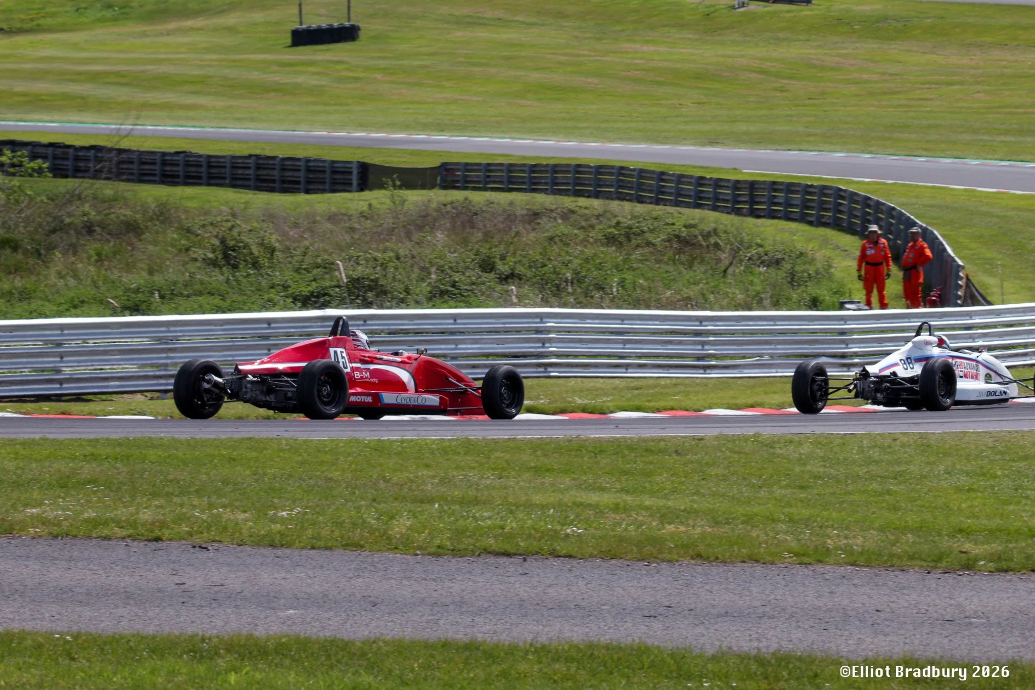 Two race cars on a racetrack with three officials in red uniforms observing from the side, green grassy hills in the background.