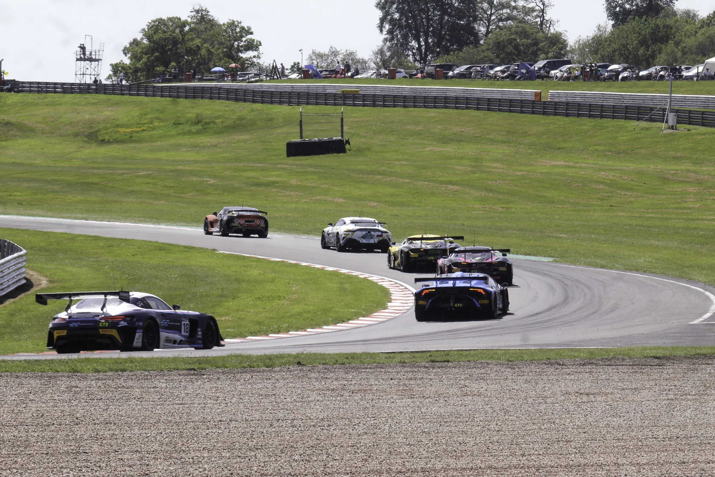 A line of racing cars on a curving race track, with spectators watching from a grassy hill, some shading with umbrellas, and parked cars in the background.