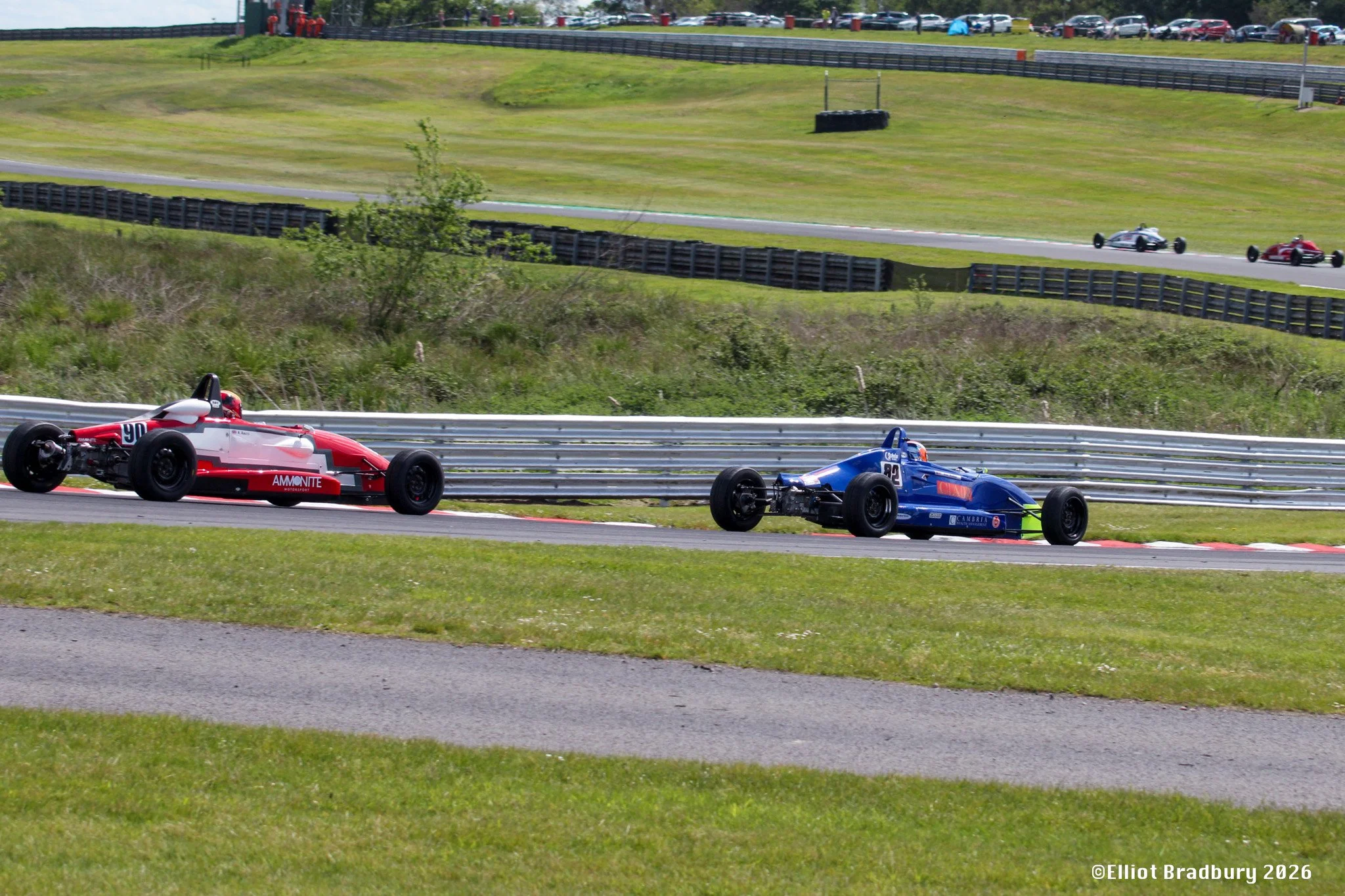 Race cars speeding on a track with spectators and cars parked in the background.
