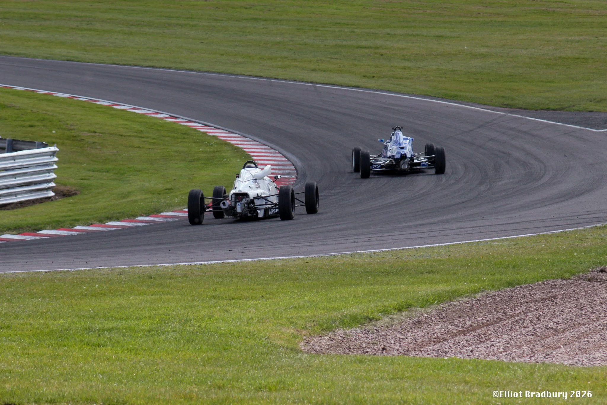 Two open-wheel race cars on a racetrack, one white and one blue, navigating a curve on a grass-lined racing circuit.