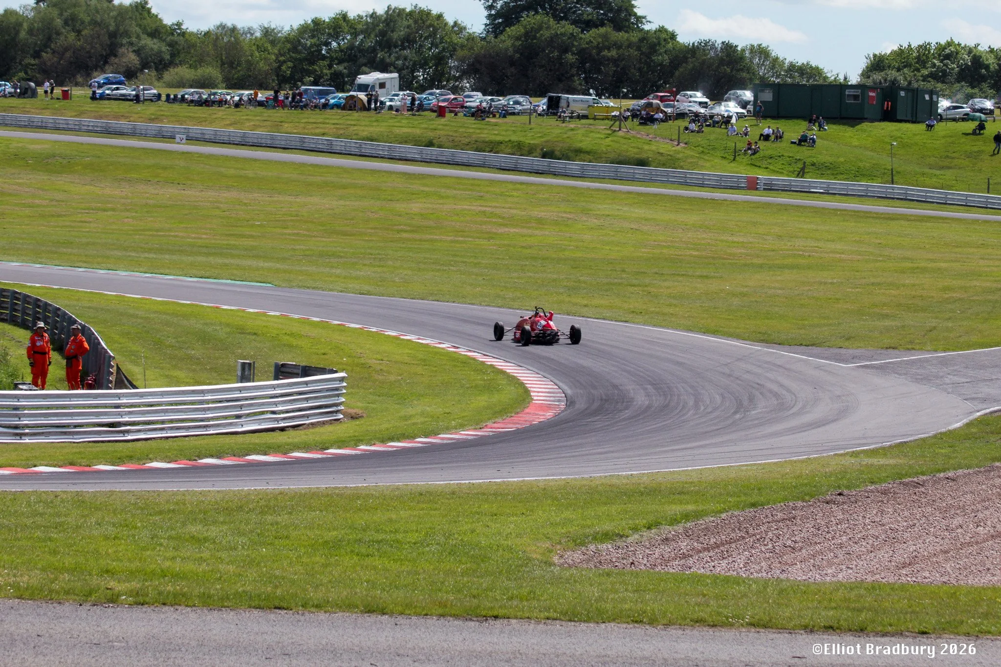 A vintage red race car on a curved section of a race track with green grass and safety barriers. In the background, there are spectators sitting on a grassy hill, cars parked, and trees under a partly cloudy sky.