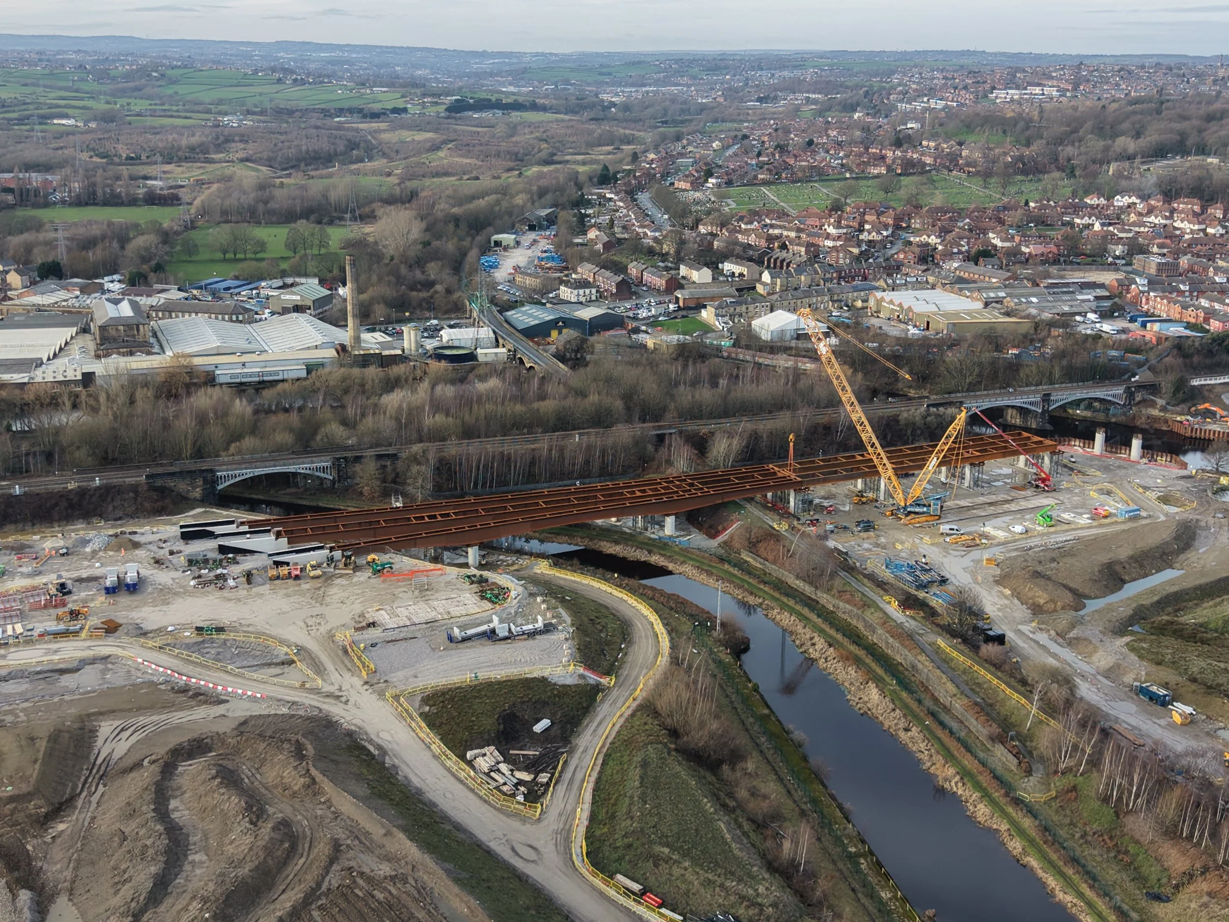 Aerial view of a construction site with cranes building a bridge over a river, surrounded by a residential area with houses and green spaces, under a cloudy sky.