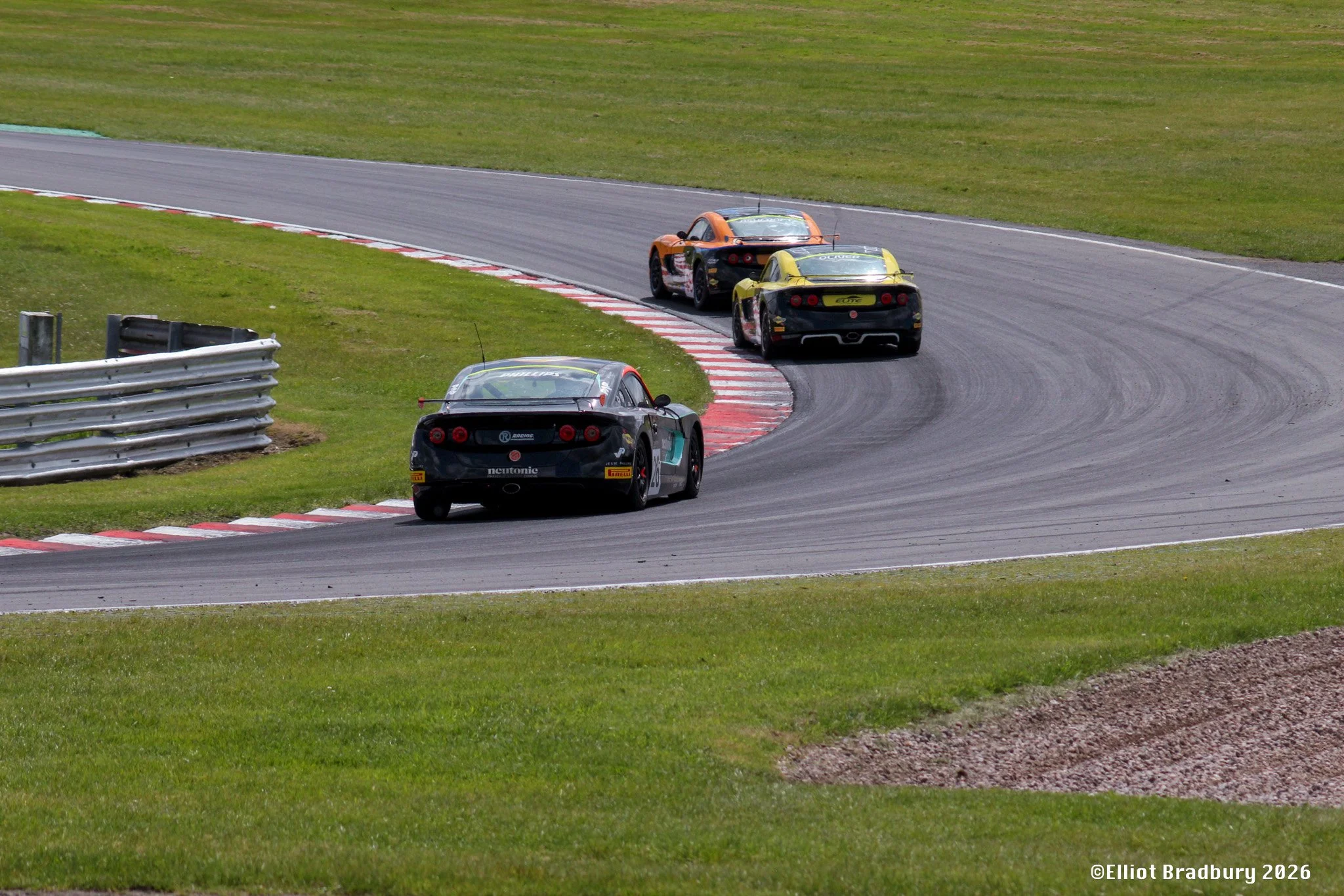 Three race cars driving on a curving race track with red and white striped curbs, metal safety barriers, and grassy surroundings.