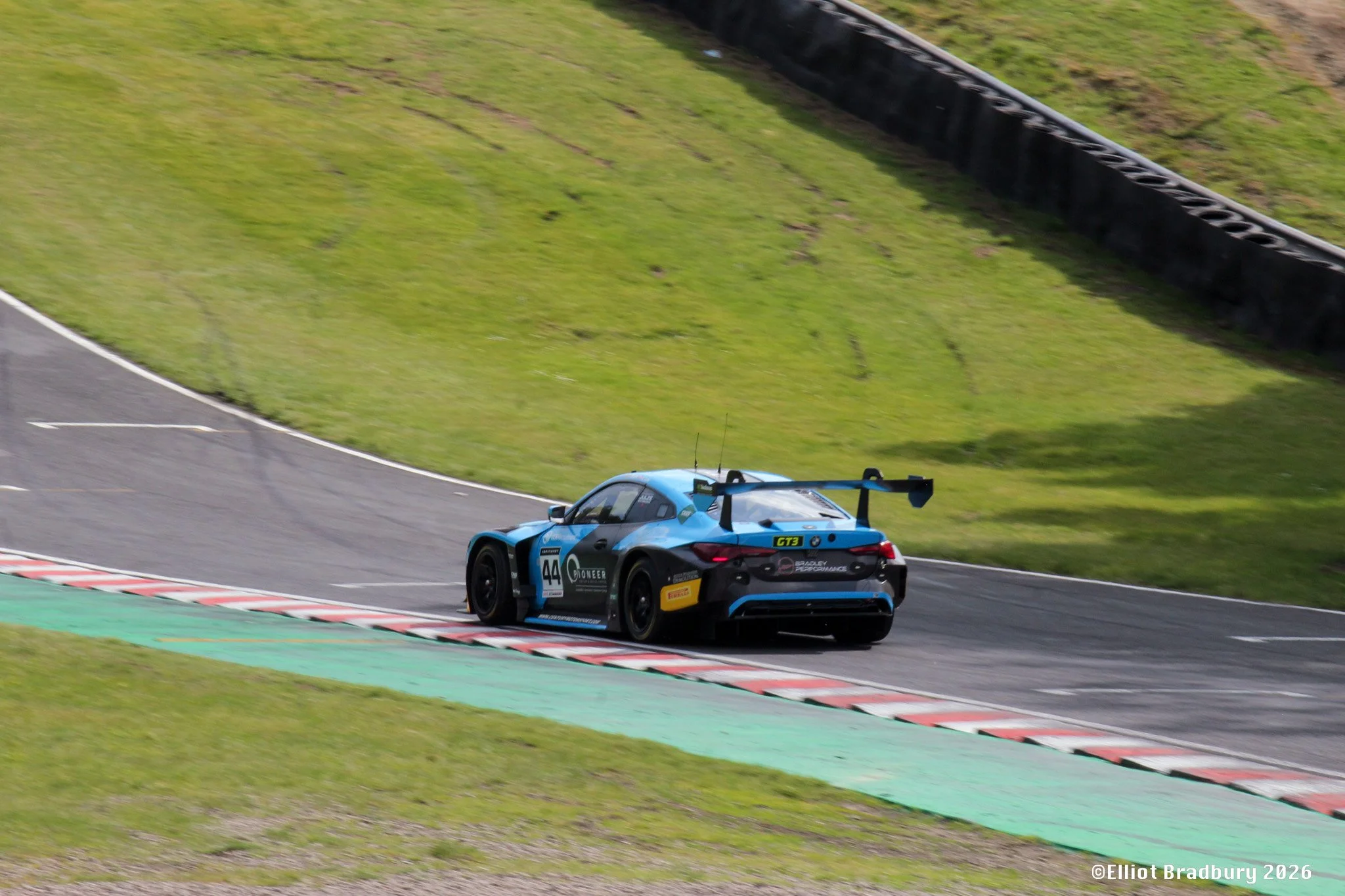 A blue race car with the number 44 on the side is driving on a racing track, approaching a corner with red, white, and green curbing, and a grassy area beside the track. A black barrier is visible along the outside of the turn.