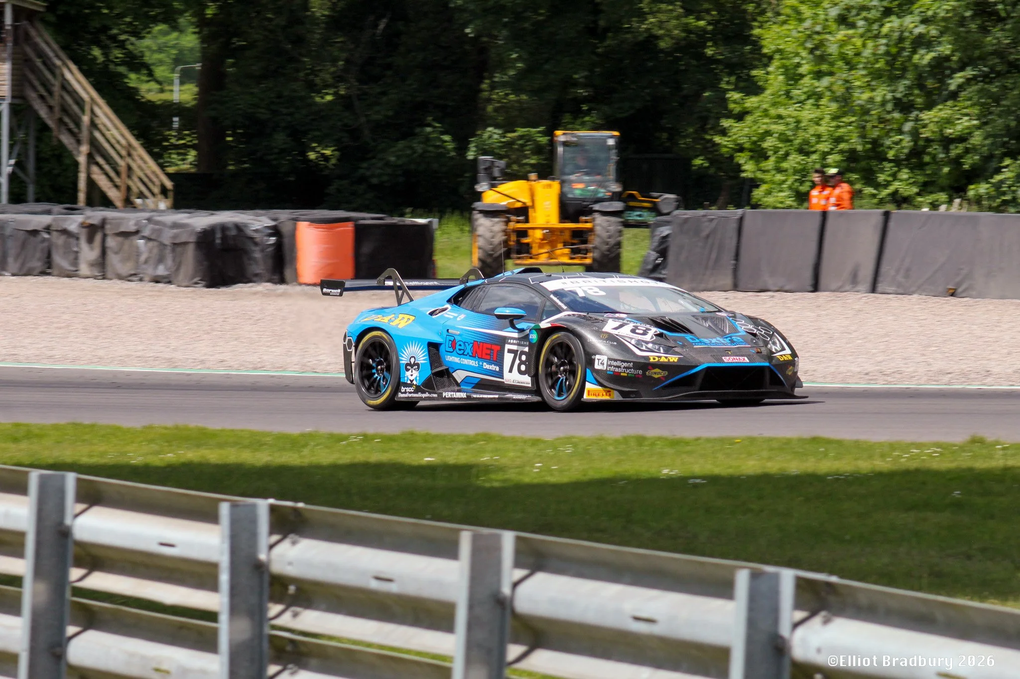 A blue race car on a race track with various sponsorship logos and the number 78, driving past safety barriers and a yellow construction vehicle, with trees and two people in orange vests in the background.