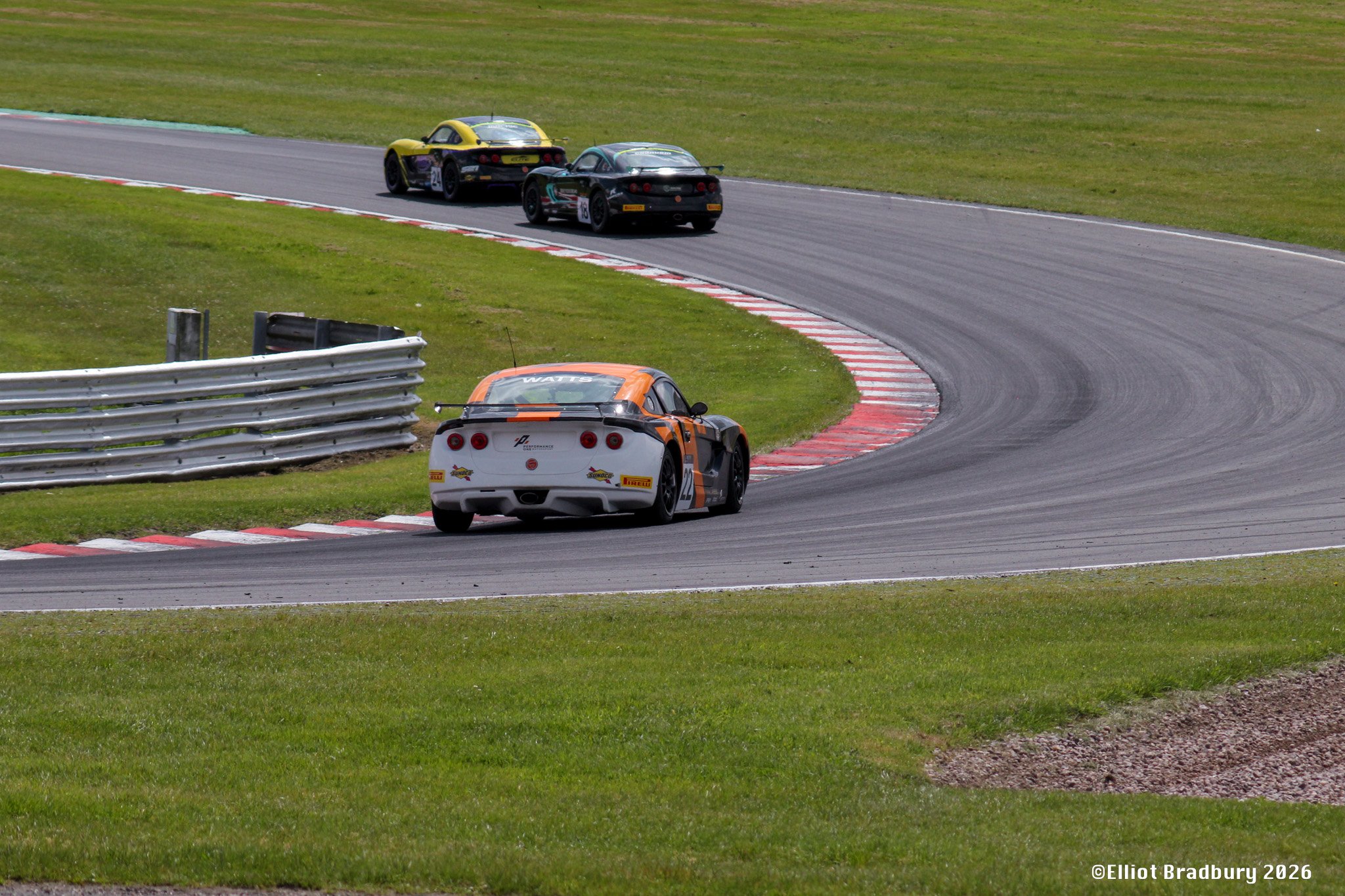 Three race cars on a racing track, navigating a curve with green grass on the sides and safety barriers.