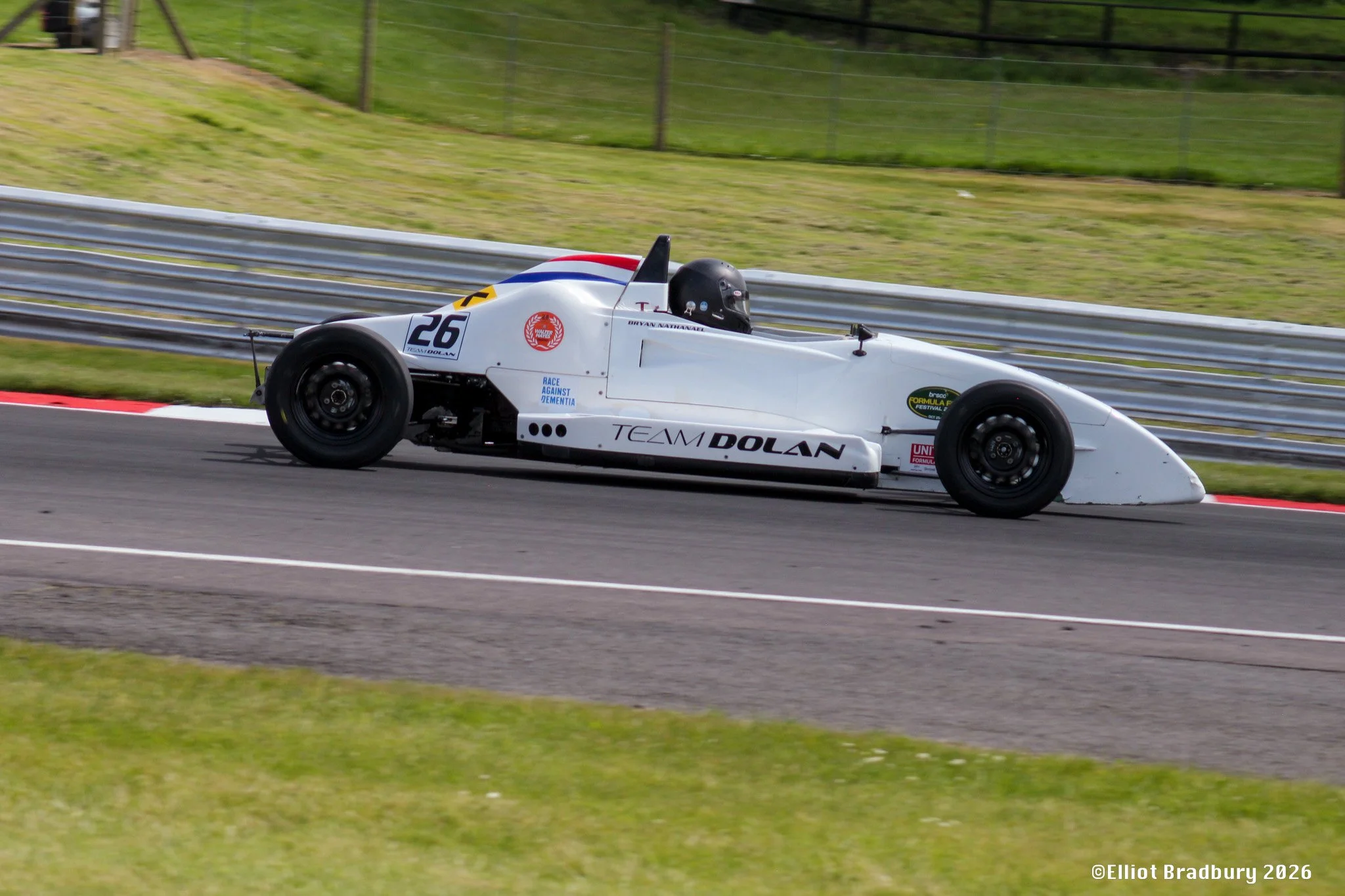 A white race car with the number 26, logo and sponsors, driven by a helmeted driver, is speeding along a racetrack with a guardrail and grassy area in the background.