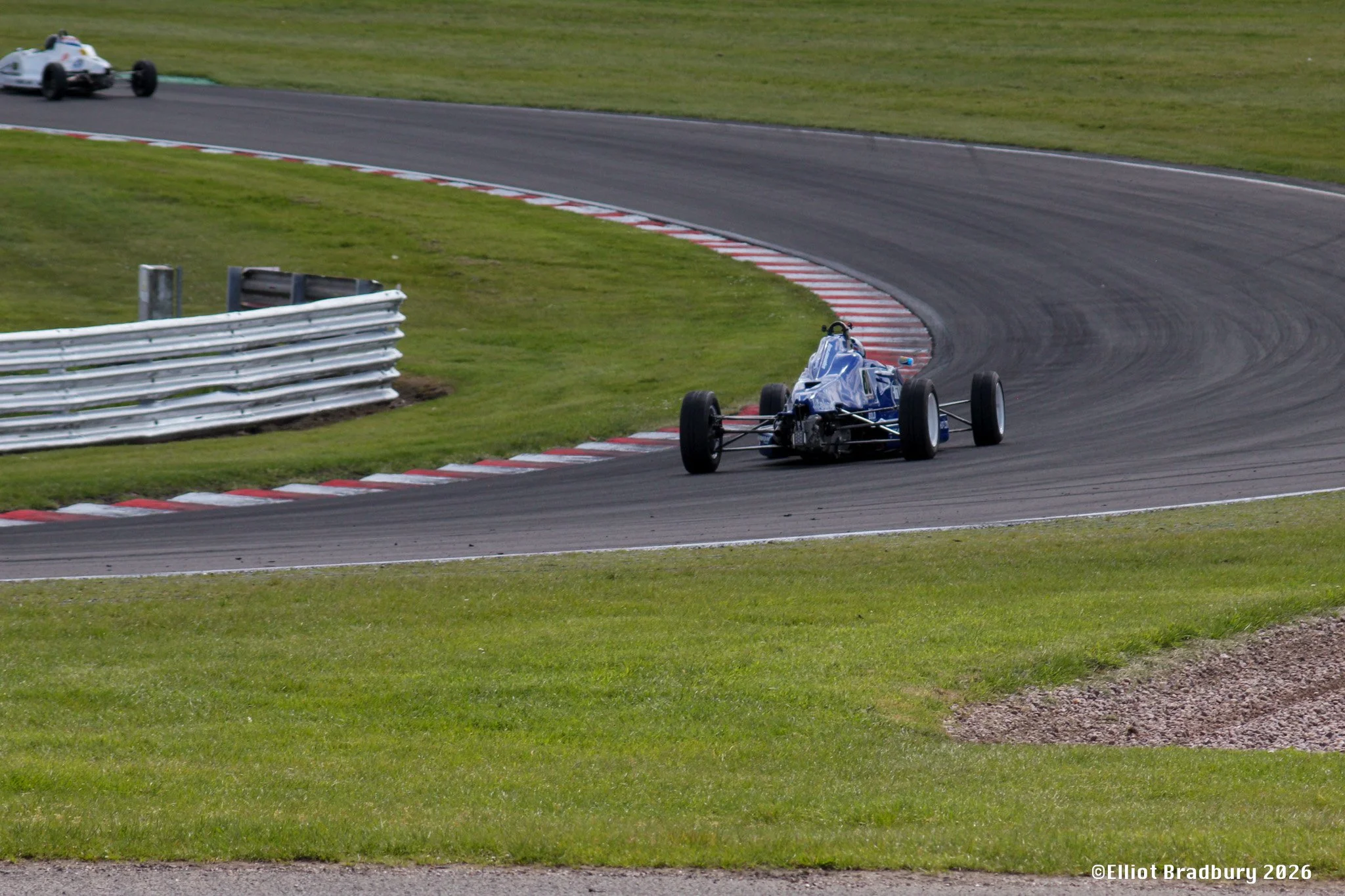A blue open-wheel race car on a racetrack taking a turn, with a white race car in the background, green grass on either side of the track, and a metal barrier on the left side.