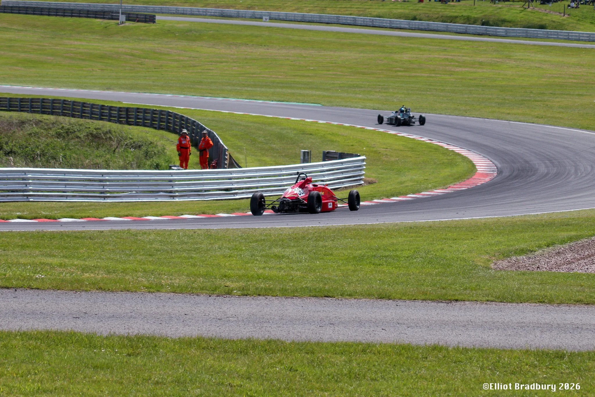 A red Formula race car on a racetrack with a black vintage race car in the distance, two officials in red jumpsuits standing behind a safety barrier, grassy areas, safety fences, and a few spectators watching from the hill.