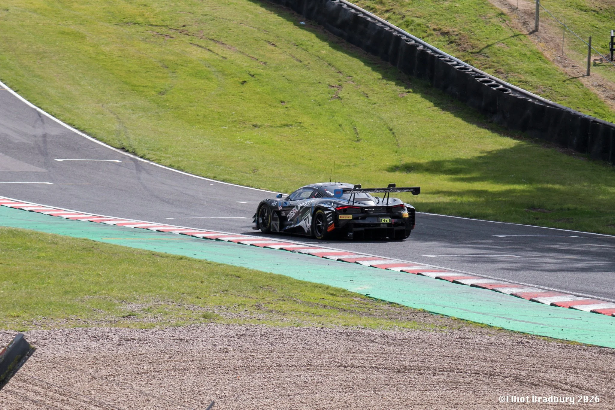 A black race car driving on a racetrack with green grass and tire barriers in the background.