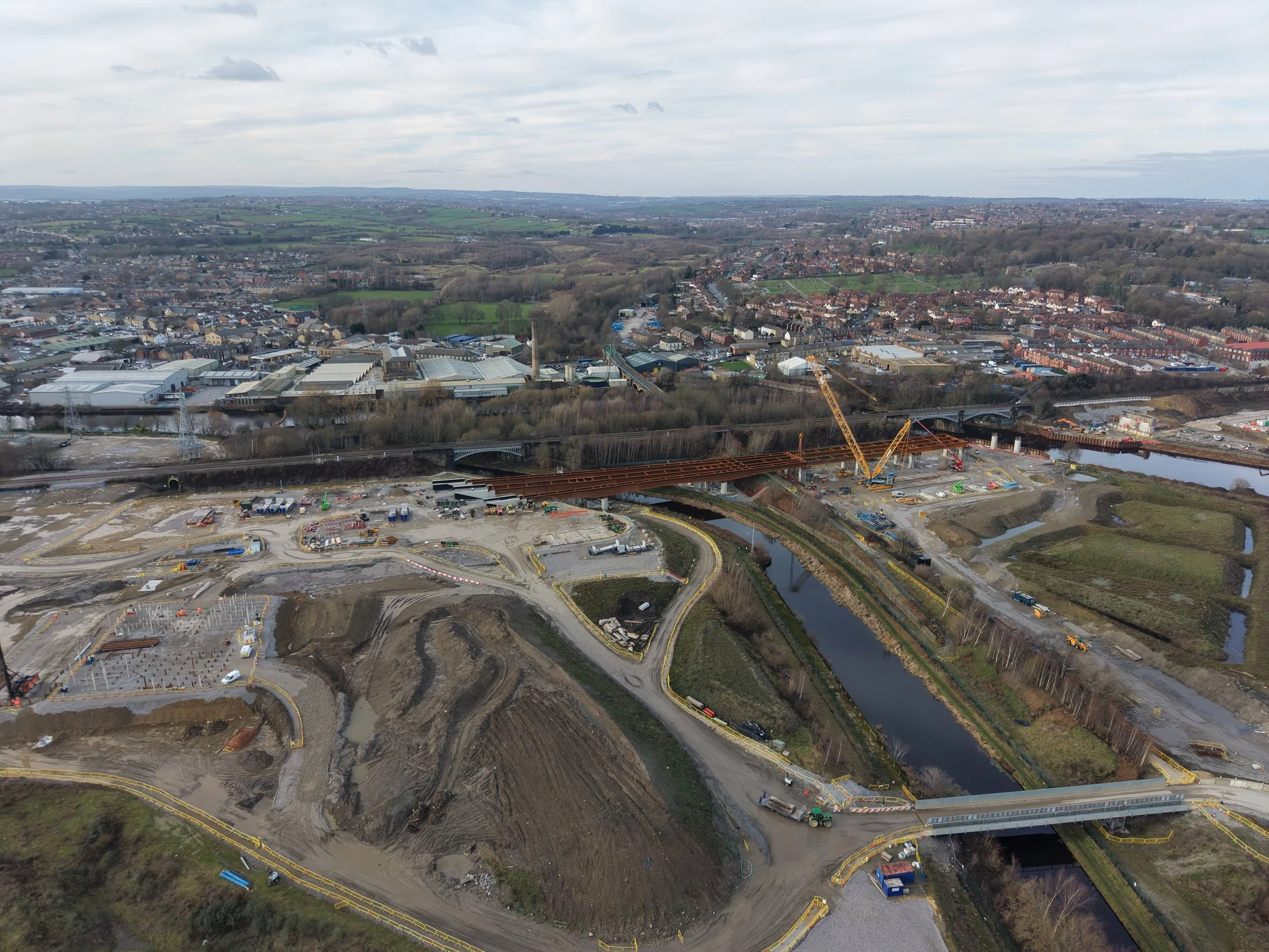 Aerial view of a cityscape with a construction site, bridges, roads, and a river, with a mix of industrial and residential areas, and green fields in the background.