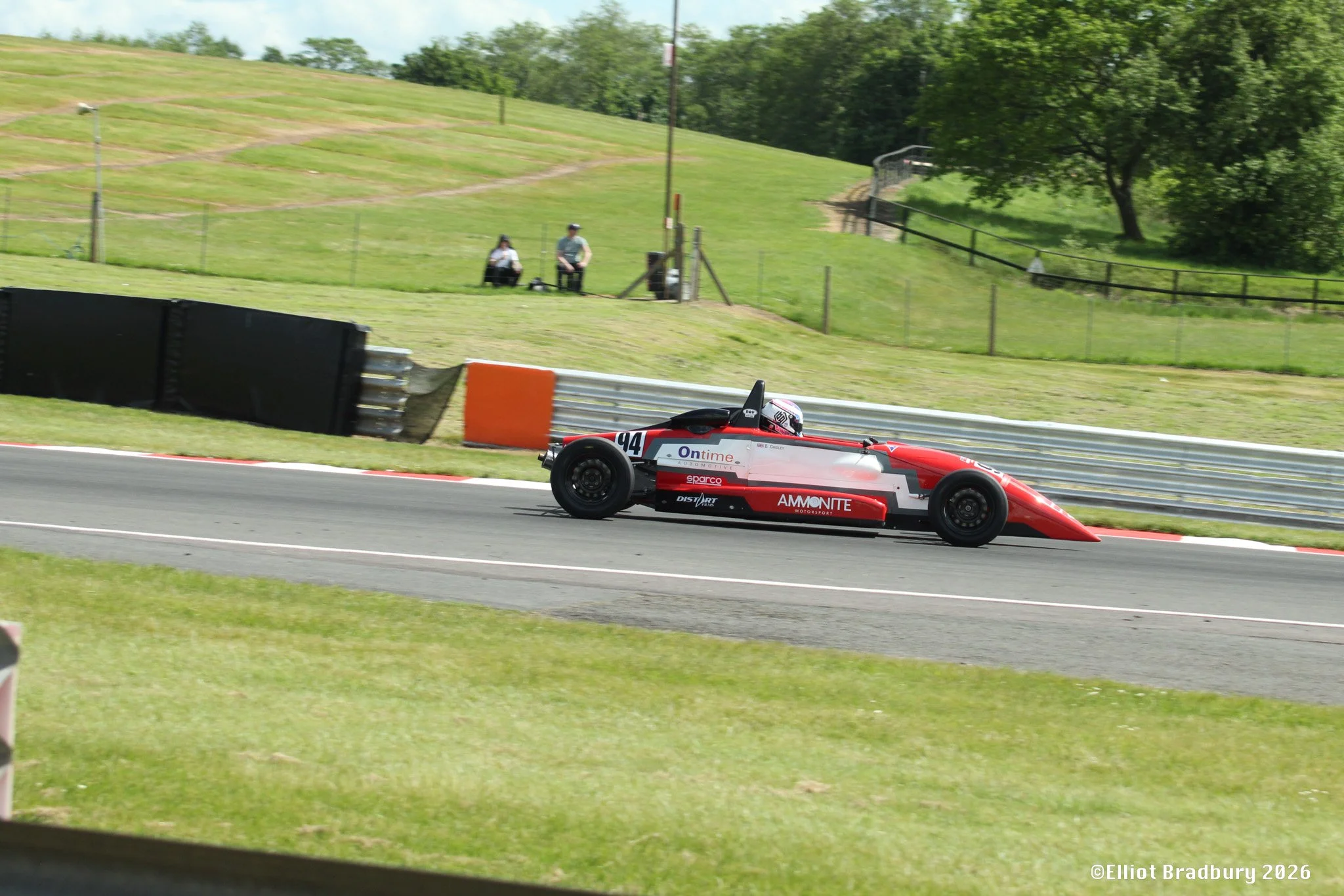 A race car with the number 94 on it driving on a race track, with grassy hills and trees in the background and two people sitting on a bench near the track.