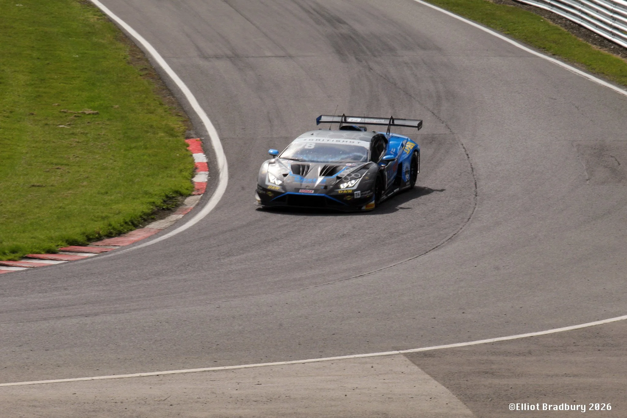A race car on a curving racing track with grassy areas and barriers, captured during a race or testing event.