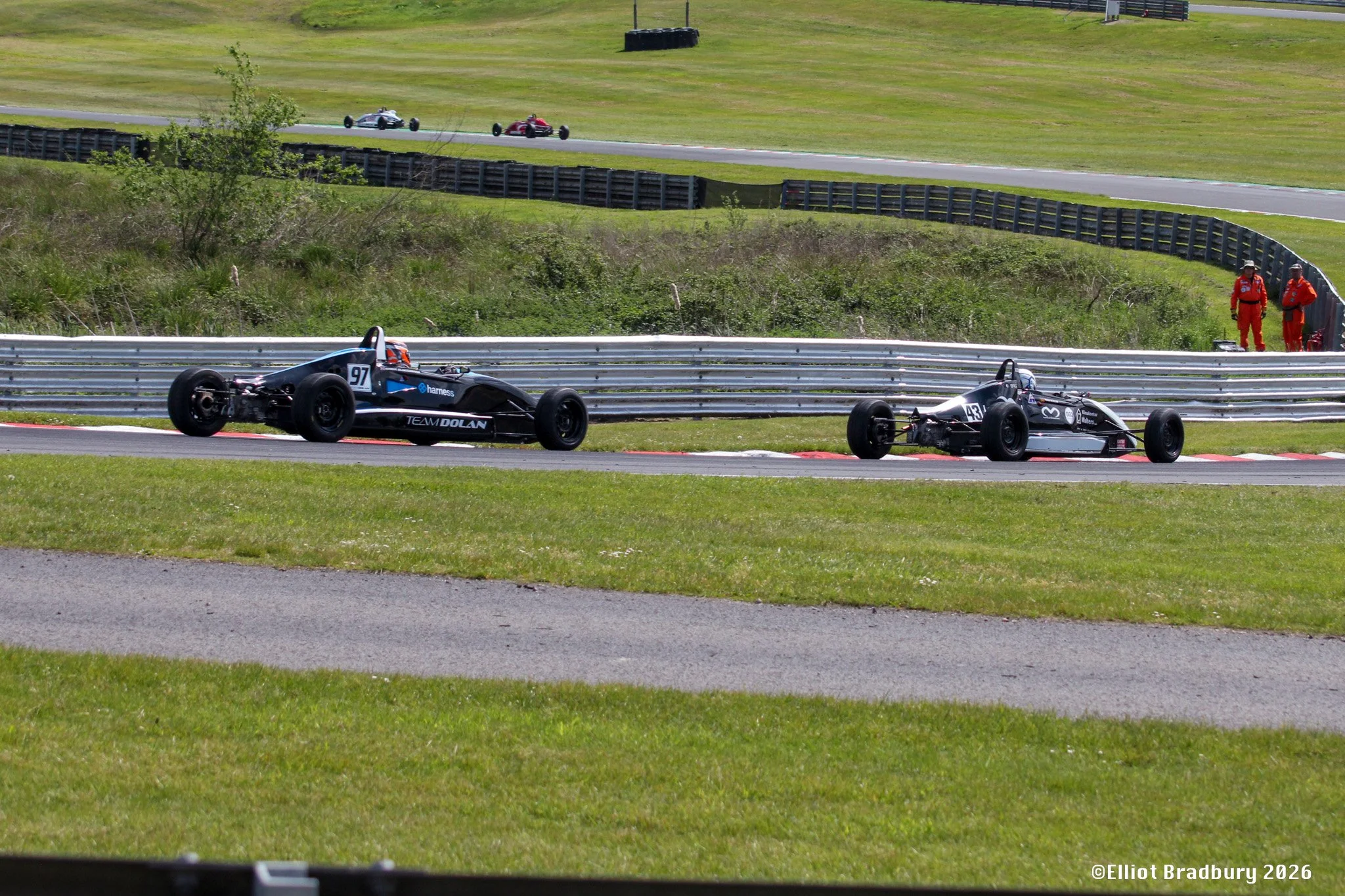 Two race cars on a track with two individuals standing by the safety barrier, surrounded by green grass and hills.