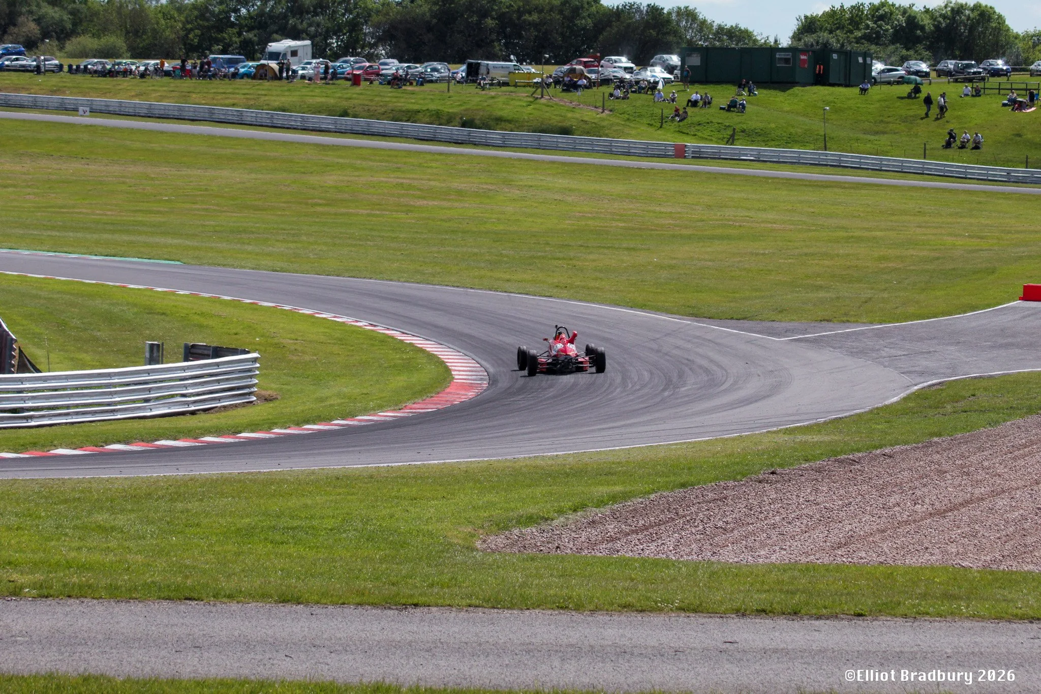 A race car on a racetrack with grassy hills and spectators in the background.