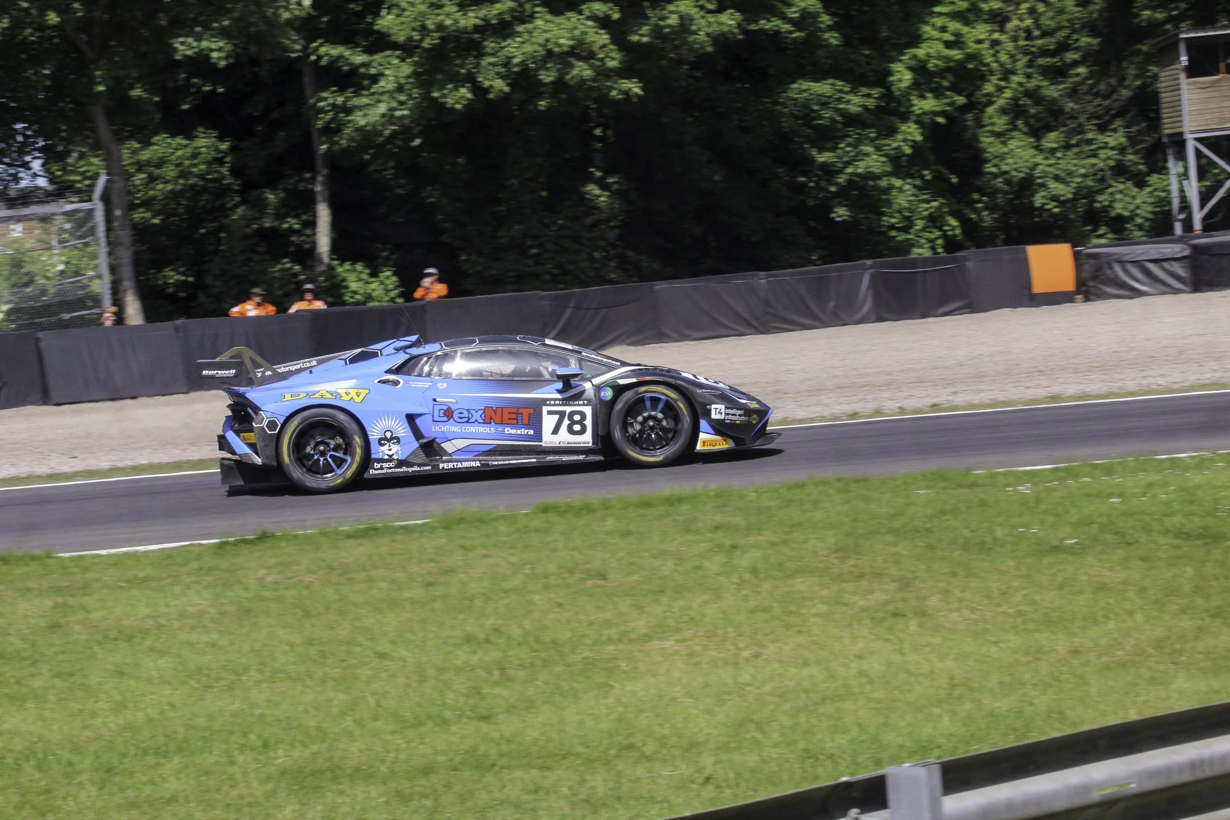 A blue racing car with the number 78 on the side, sponsored by DexNet, driving on a race track with trees and caution barriers in the background.